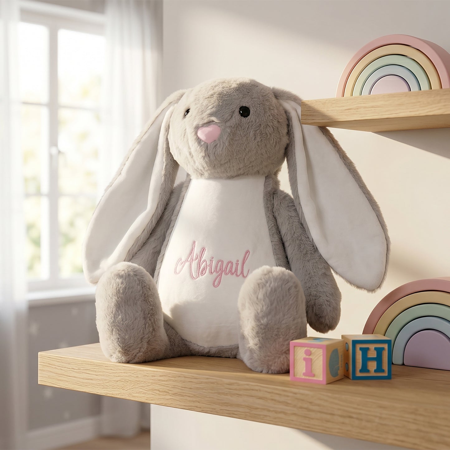 A plush gray bunny stuffed animal with long ears sits on a wooden shelf in a softly lit room. The center of the bunny is white with the name 'Abigail' stitched in pink script.