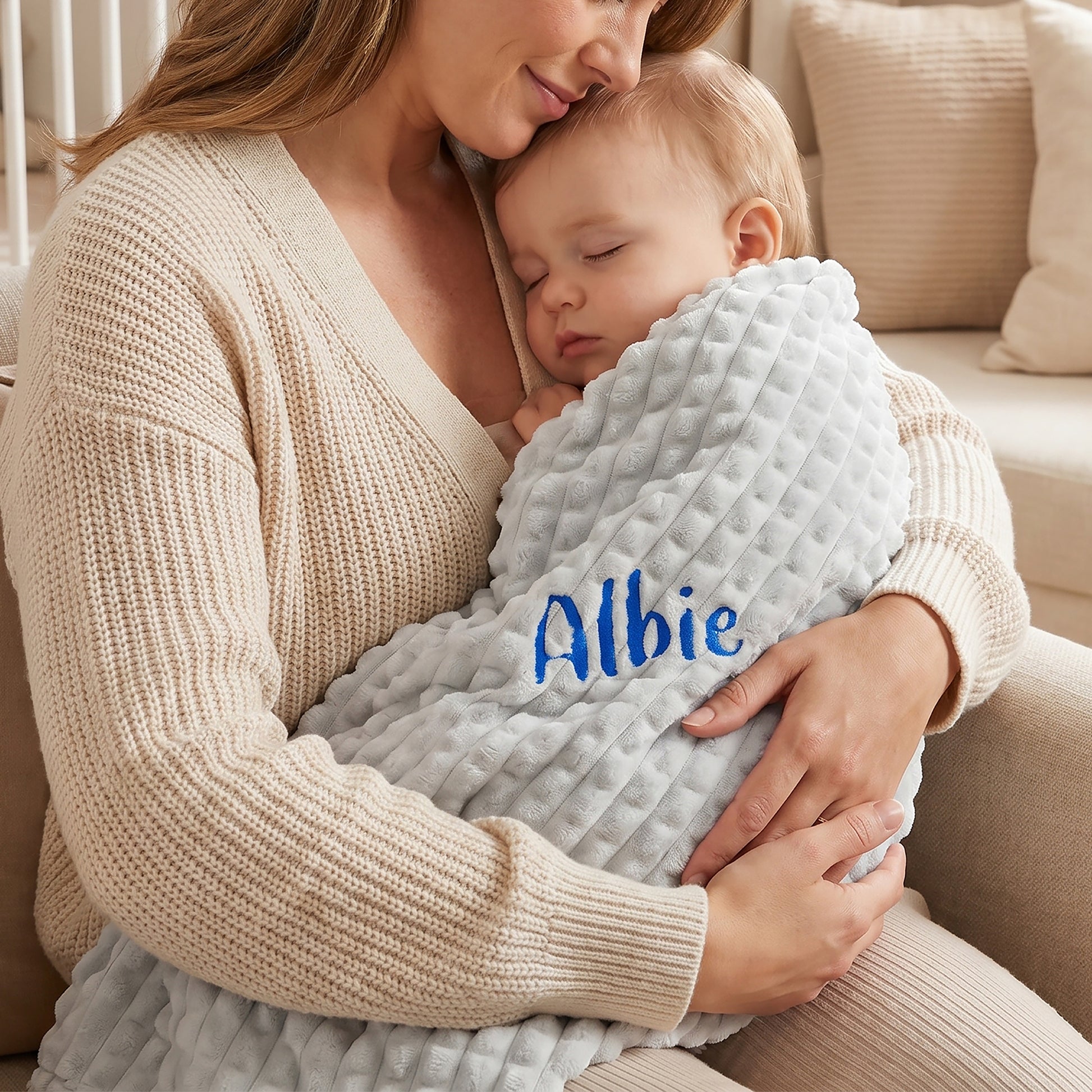 A mother in a cream knit sweater holds her sleeping baby, who is wrapped in a light gray minky dot blanket with the name 'Albie' embroidered in blue on the front, set in a cozy living room.