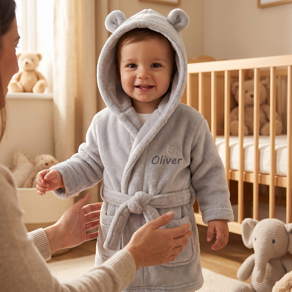 A baby wearing a light gray plush hooded bathrobe with bear ears and the name 'Oliver' embroidered on the chest, standing in a softly lit nursery while an adult's hands reach out to help.