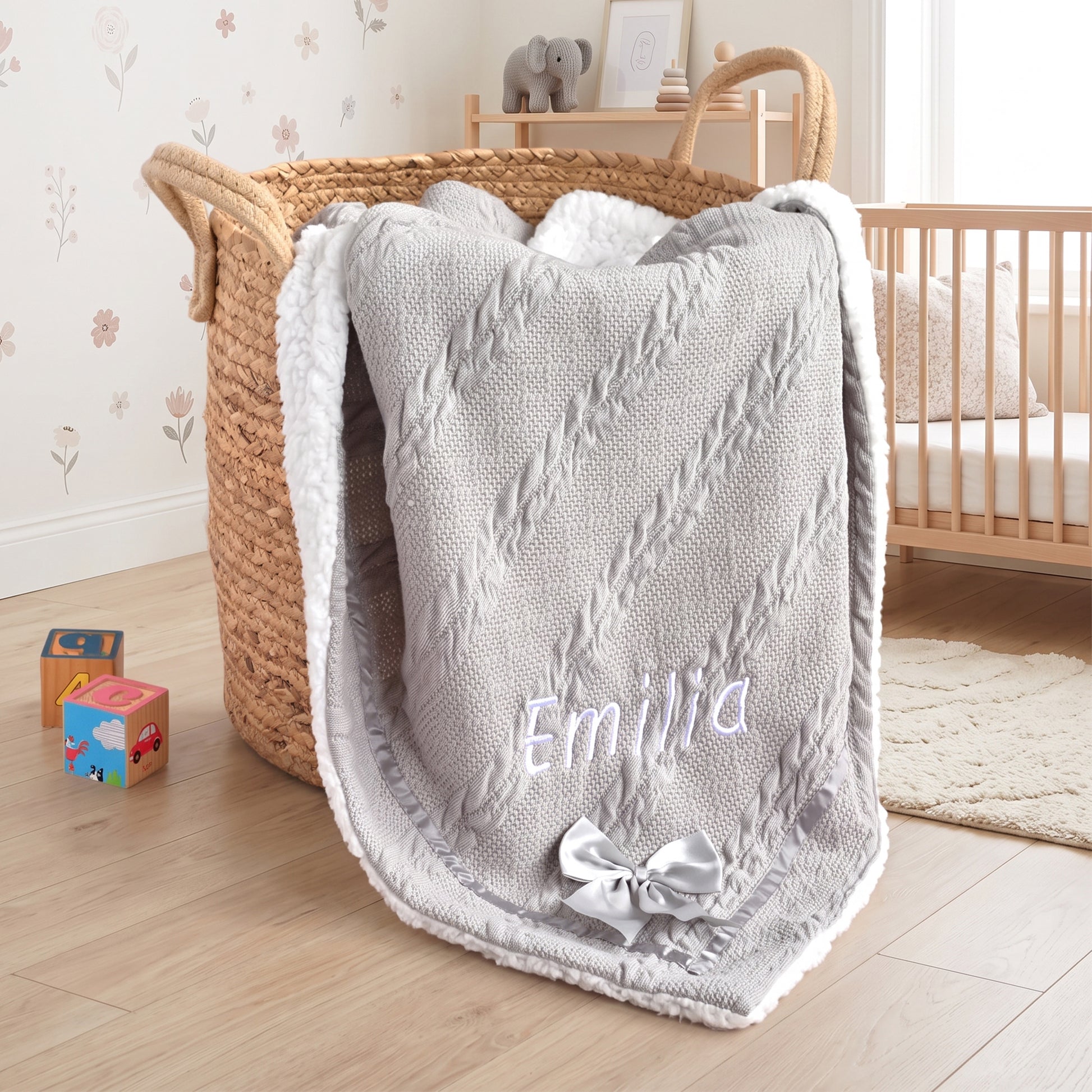 Grey cable-knit baby blanket with sherpa fleece backing and satin ribbon border, personalised with the name Emilia embroidered in white, finished with a grey satin bow.