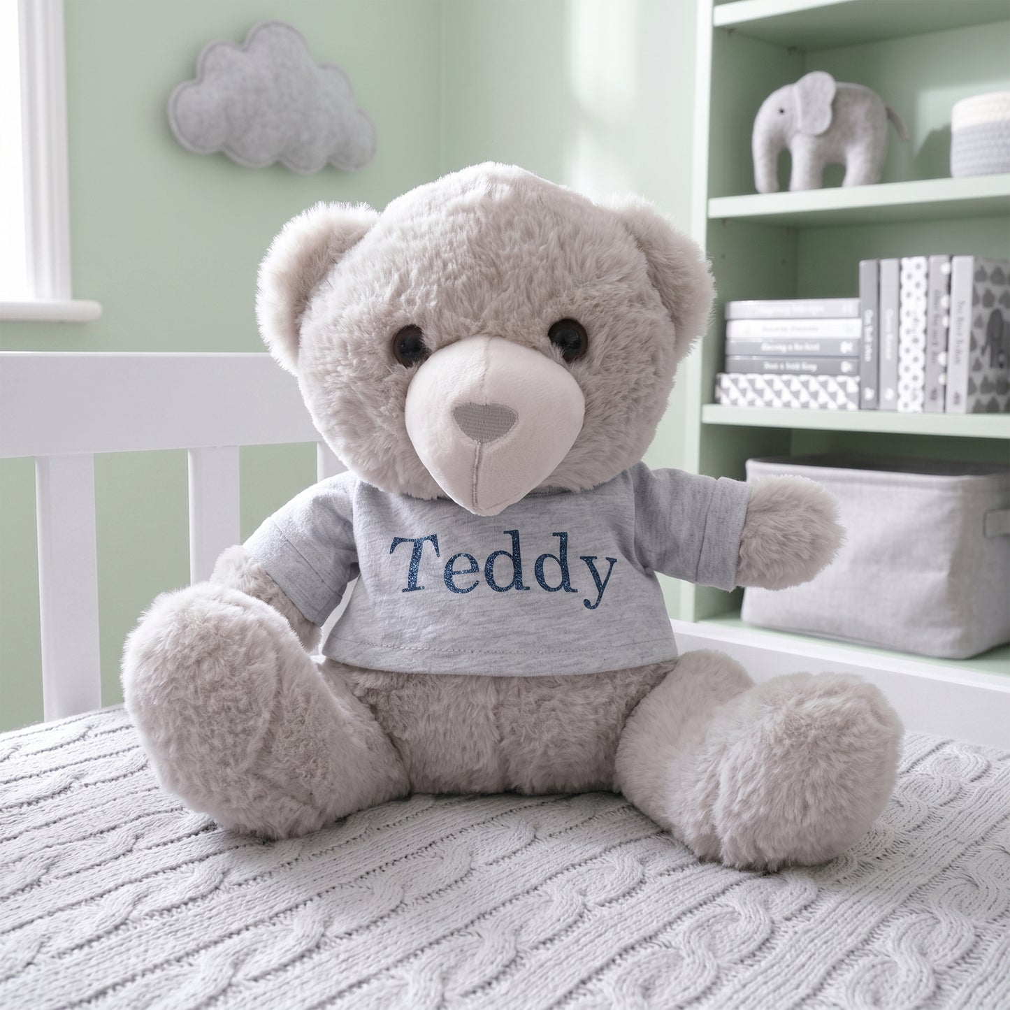 A plush light gray teddy bear sits on a white bed in a softly lit nursery. The bear wears a light gray T-shirt with the name 'Teddy' printed in navy blue.