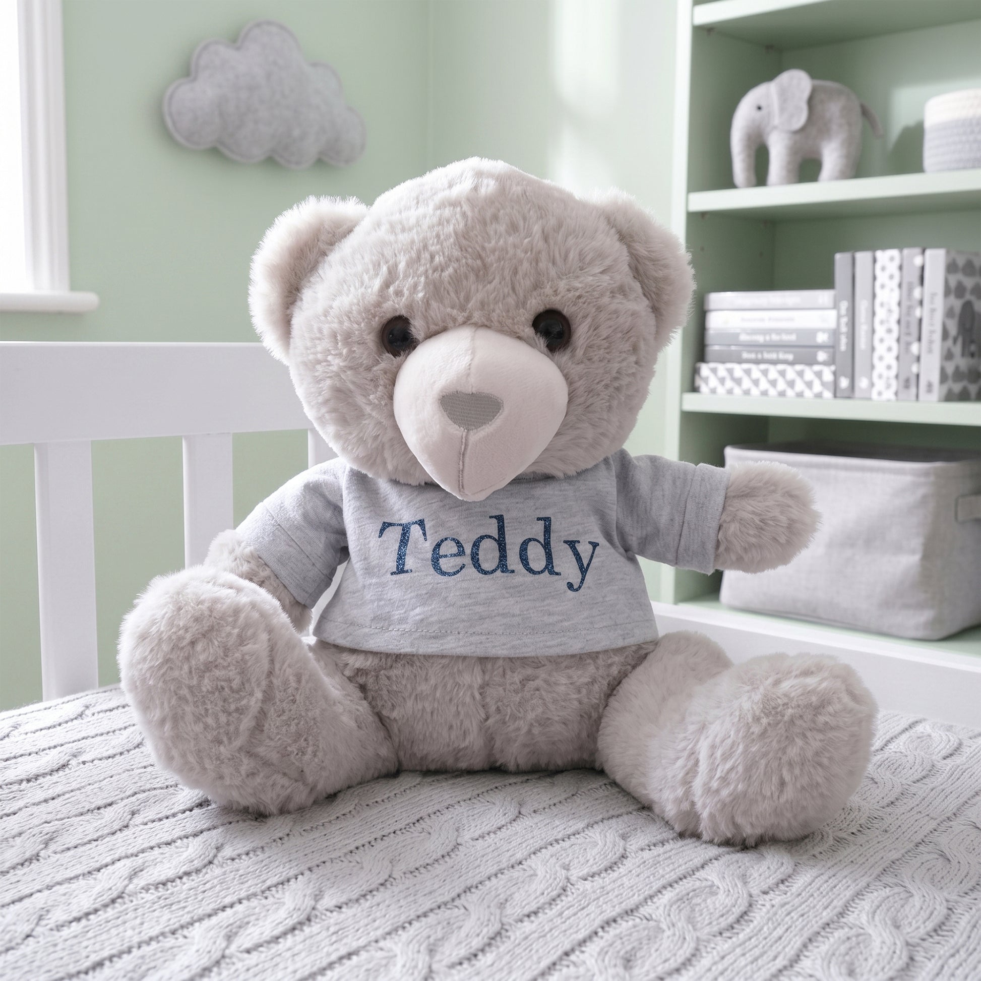 A plush light gray teddy bear sits on a white bed in a softly lit nursery. The bear wears a light gray T-shirt with the name 'Teddy' printed in navy blue.