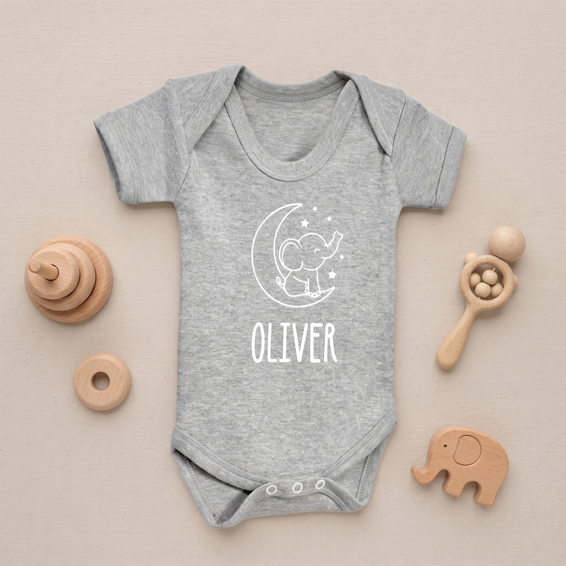 A heather gray short-sleeve baby bodysuit with a white crescent moon and elephant graphic and the name 'OLIVER' printed on the front, styled in a flat lay on a beige background with wooden baby toys.