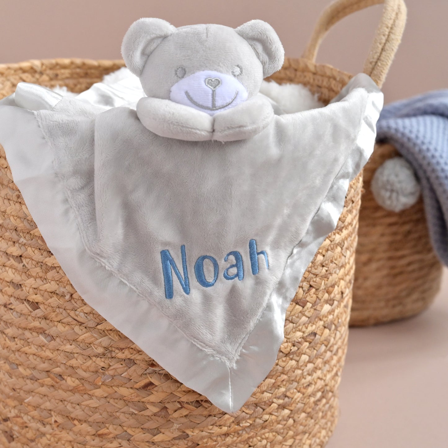 A personalized baby security blanket featuring a plush gray teddy bear head and a white satin fabric body with the name 'Noah' embroidered in blue. The blanket is draped inside a woven straw basket.