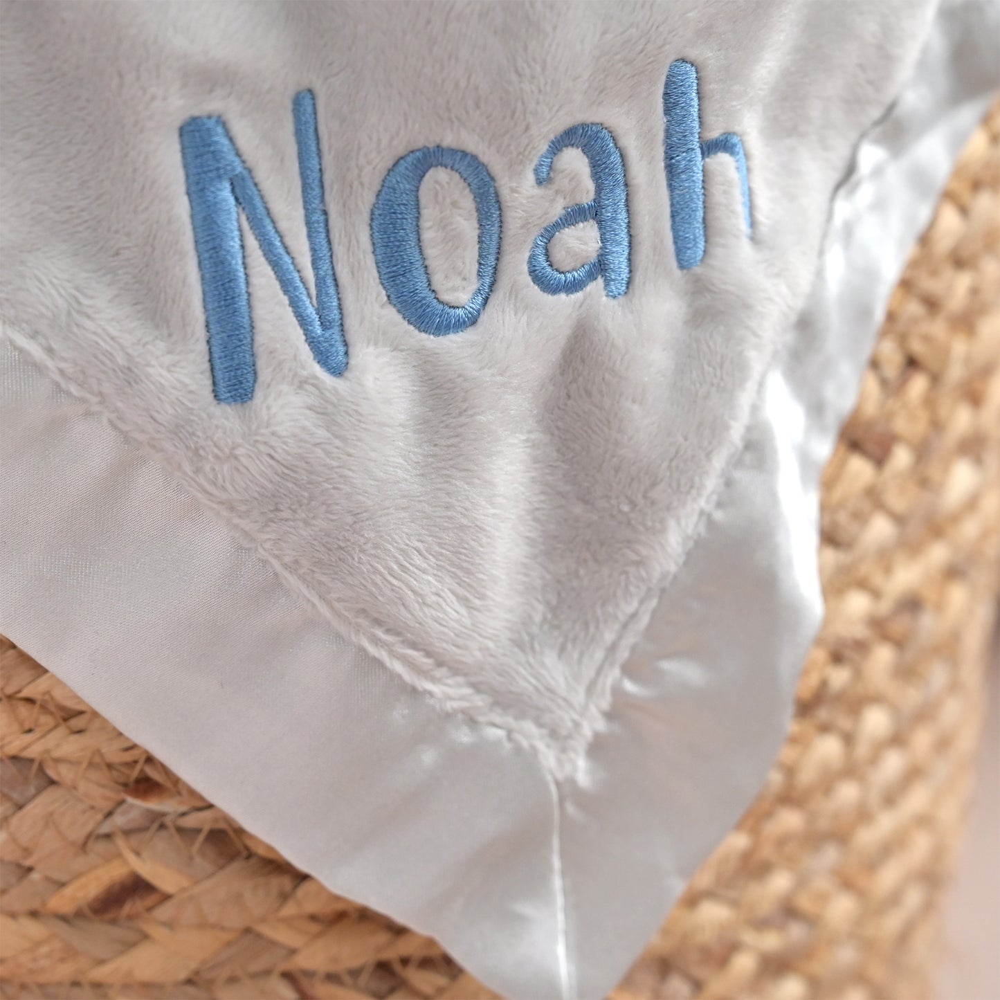 Close-up of a white plush baby blanket with light blue embroidered name 'Noah' and a satin border, resting on a woven straw surface.