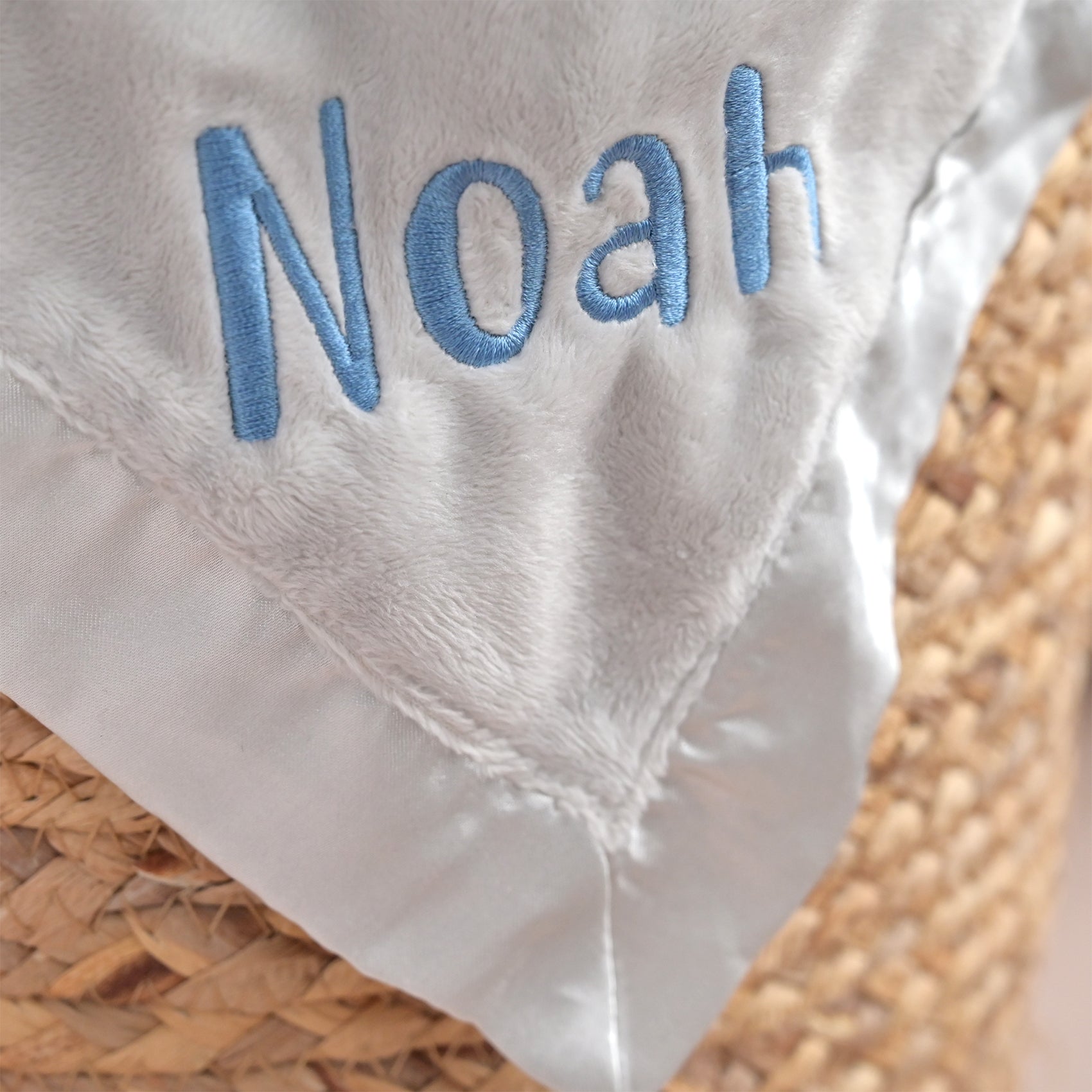 Close-up of a white plush baby blanket with light blue embroidered name 'Noah' and a satin border, resting on a woven straw surface.