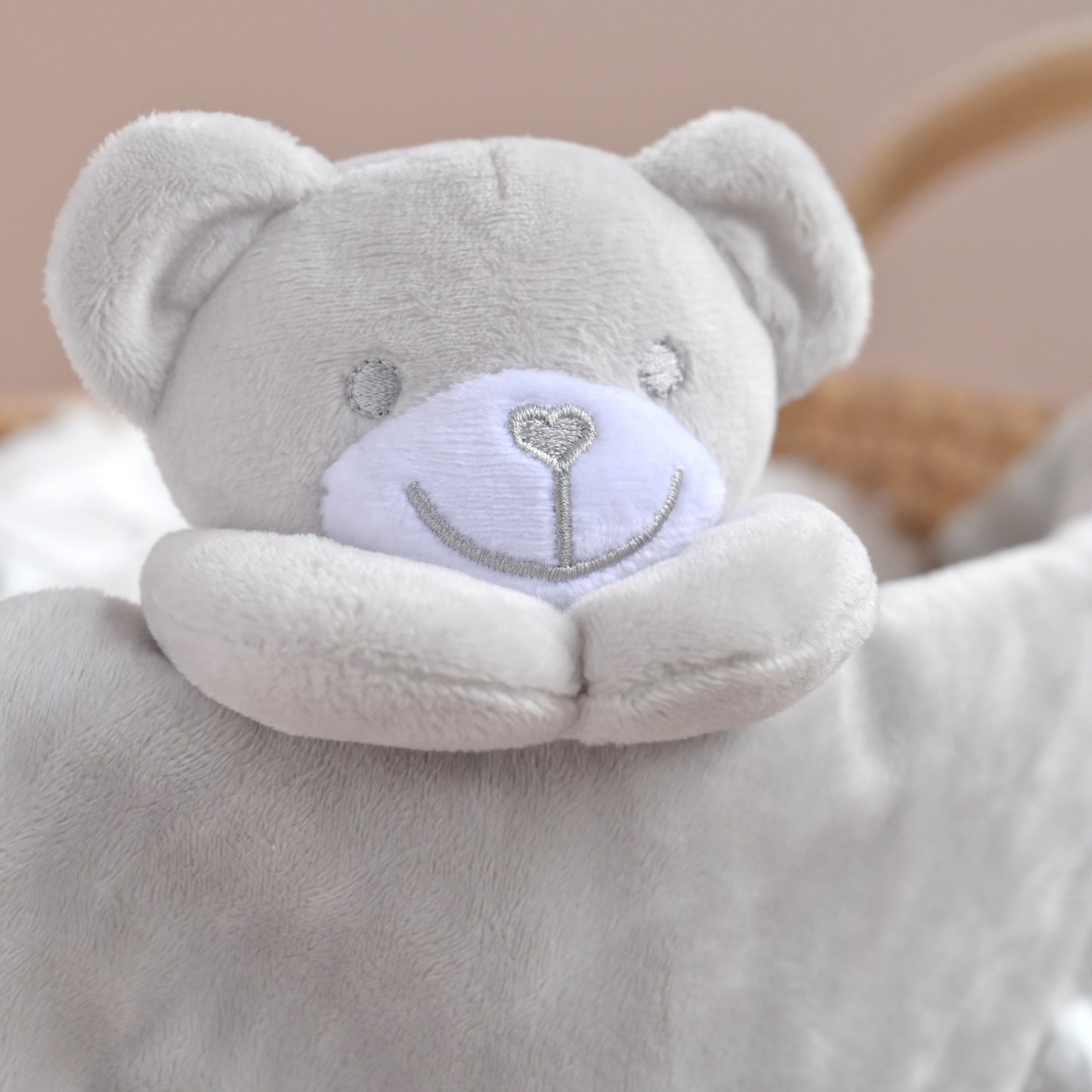 A close-up of a soft plush bear comforter in light gray with a pale lavender embroidered face and silver facial details, resting in a soft neutral setting.