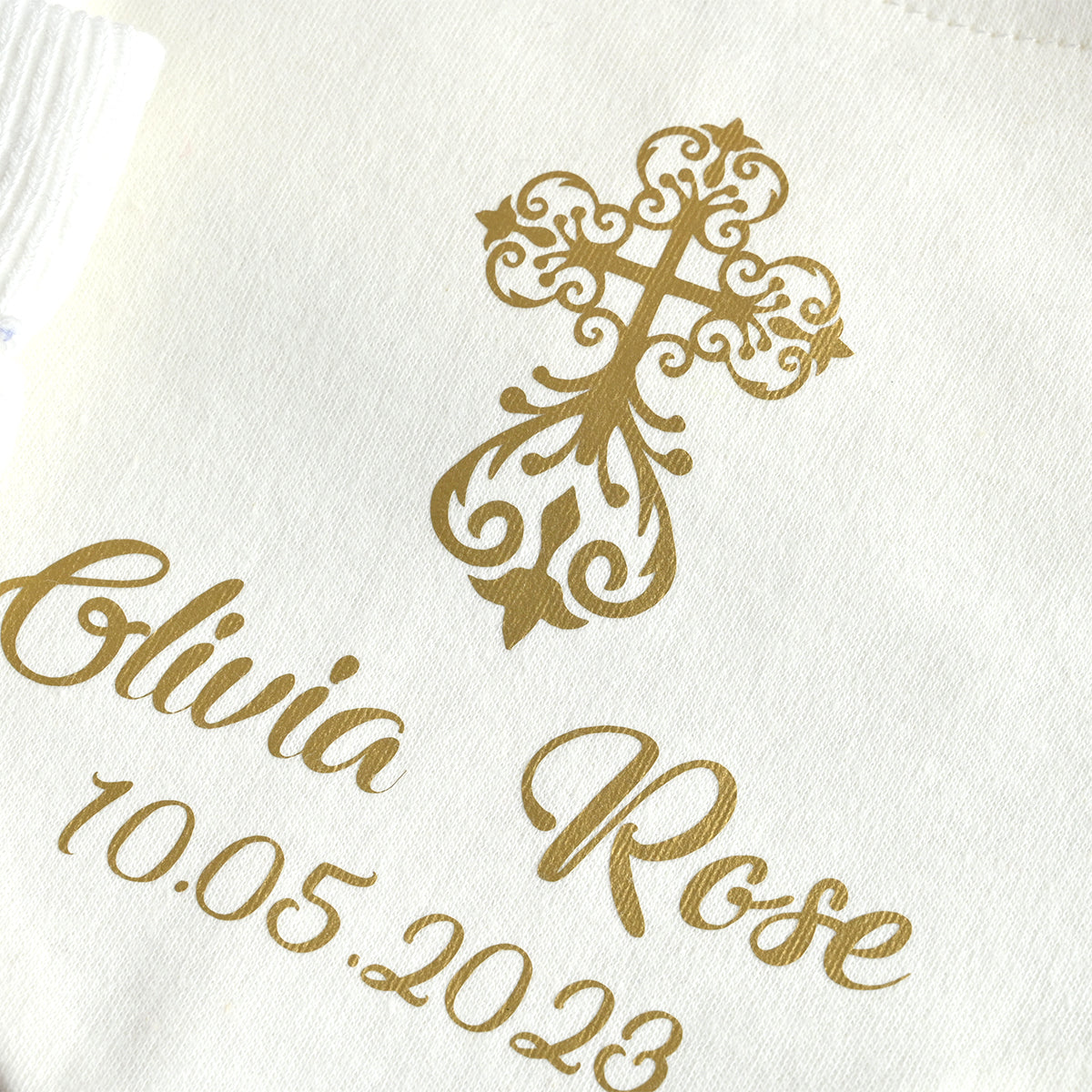 Close-up of a white fabric item featuring a gold ornate cross design and personalized text reading 'Olivia Rose 10.05.