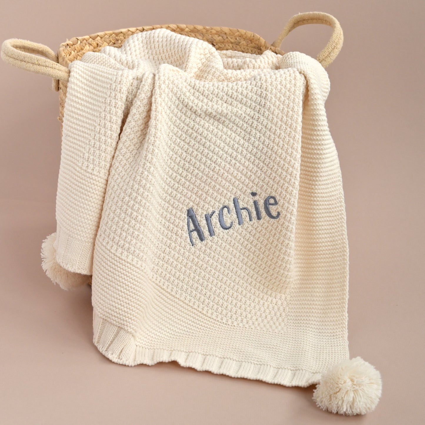 A cream-colored textured knit baby blanket with a pom-pom and the name 'Archie' embroidered in blue, draped over a woven basket against a solid beige background.