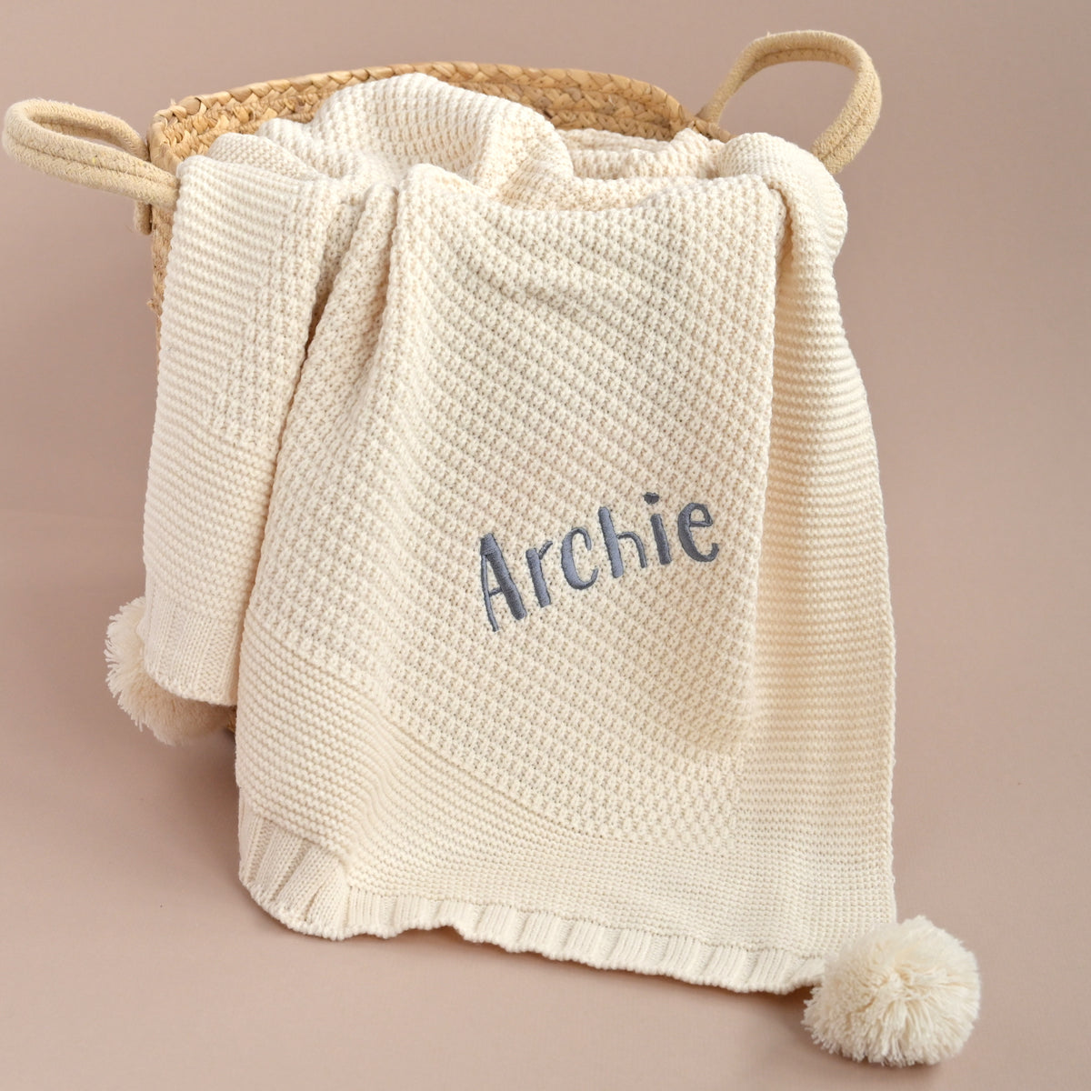 A cream-colored textured knit baby blanket with a pom-pom and the name 'Archie' embroidered in blue, draped over a woven basket against a solid beige background.