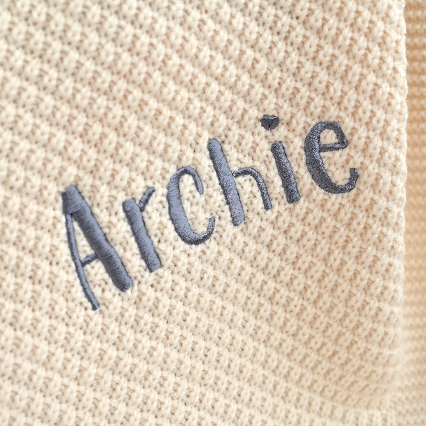 Close-up of a beige knit fabric with the name 'Archie' embroidered in dark gray lettering.
