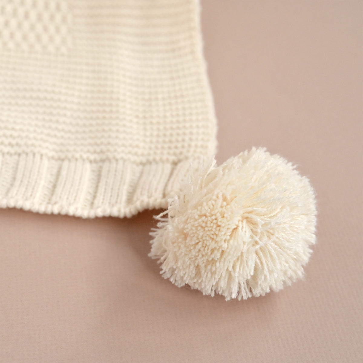 Close-up of a cream knit textile with a matching fluffy pom-pom on a soft beige background.