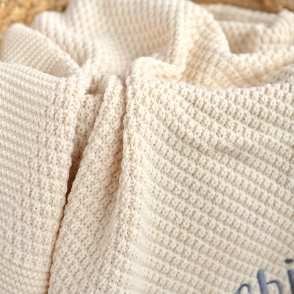 Close-up of a cream-colored textured knit blanket with a waffle or basketweave pattern, draped in soft folds.