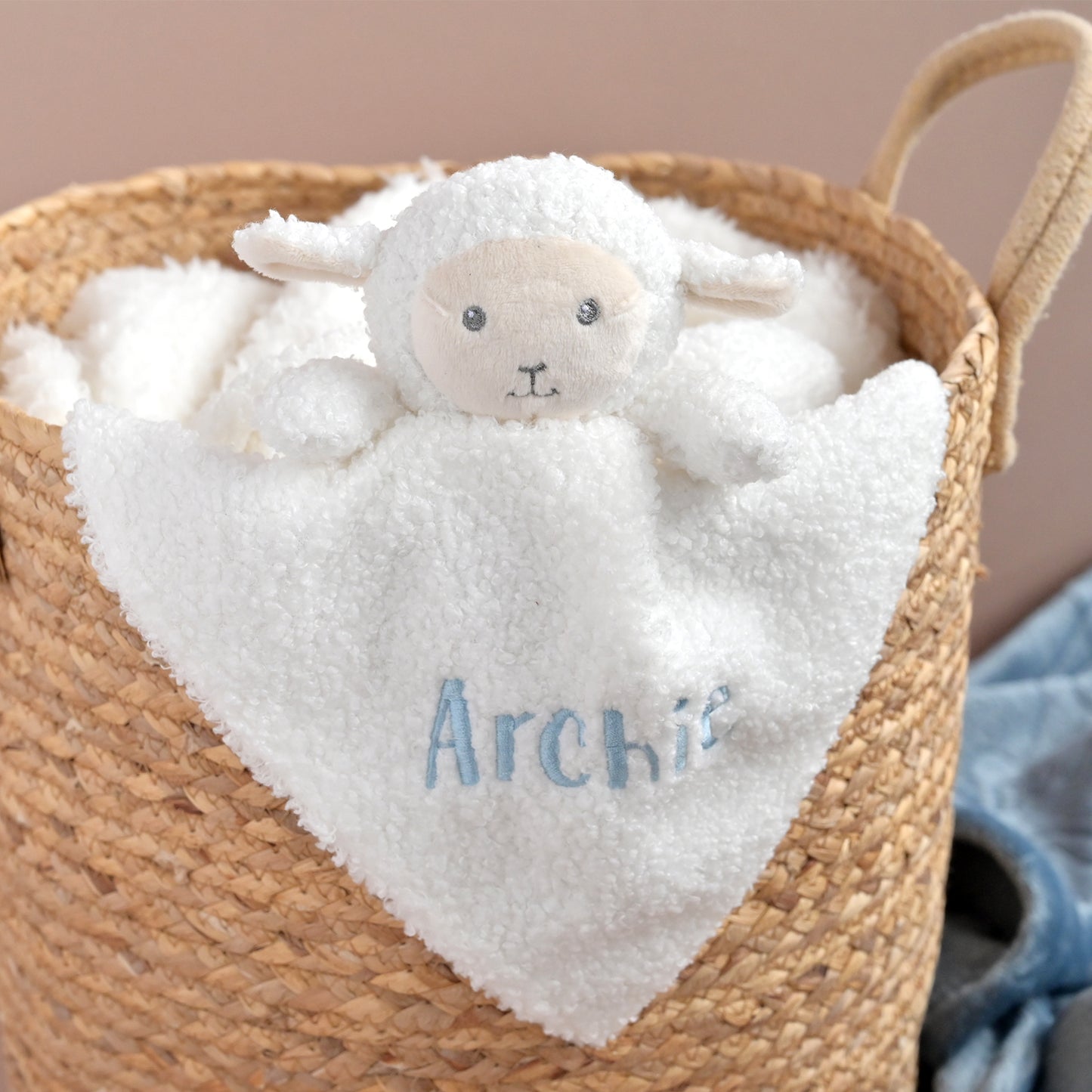 A white plush sheep security blanket with a soft face and embroidered name 'Archie' in blue, tucked inside a woven straw basket.