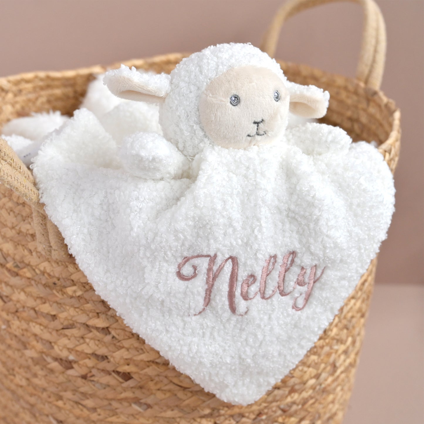 A white plush sheep security blanket with a soft heart-shaped body and embroidered name 'Nelly' in dusty pink, resting inside a woven straw basket.