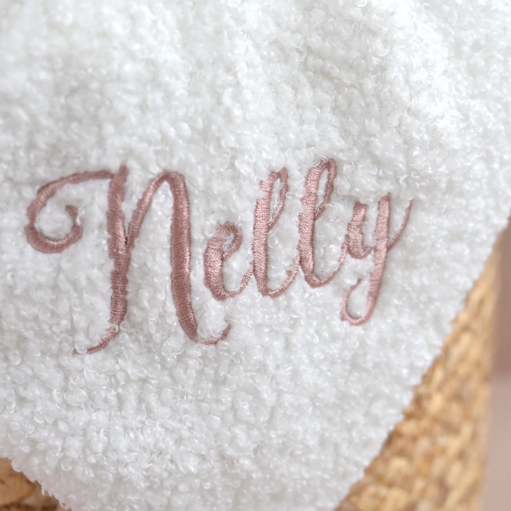 A close-up of a white sherpa or fleece blanket with the name 'Nelly' embroidered in dusty rose cursive script, resting on a woven straw basket.