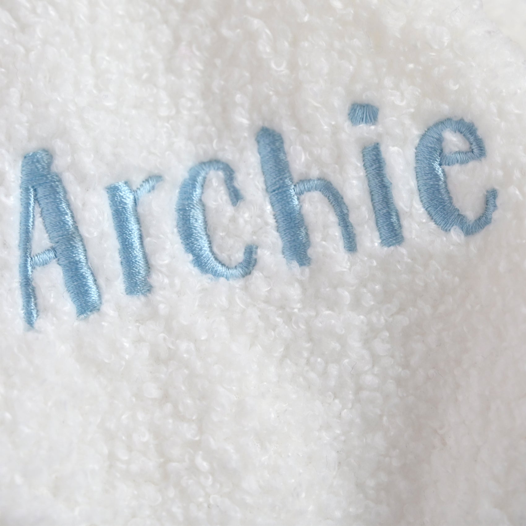 A close-up of a white sherpa fleece blanket with the name 'Archie' embroidered in light blue thread.
