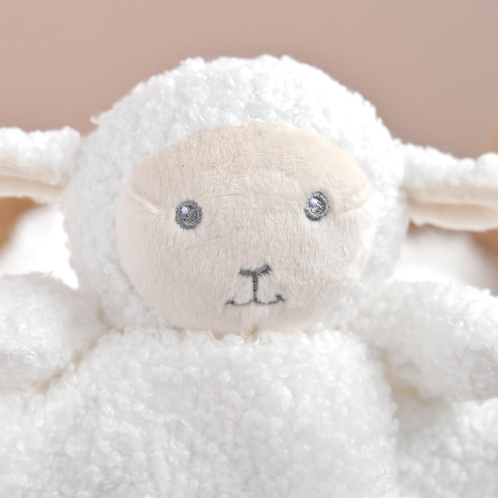 Close-up of a white plush sheep toy with a soft cream-colored face, embroidered black nose and mouth, and shiny silver bead eyes, set against a warm beige background.