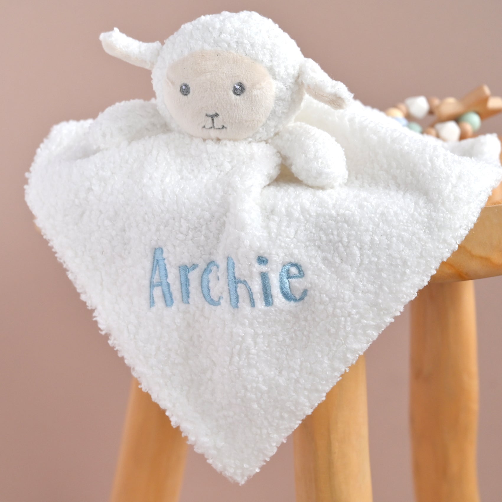 A white plush sheep lovey blanket with a soft teddy face, draped over a wooden stool. The front of the blanket is personalized with the name 'Archie' embroidered in light blue.