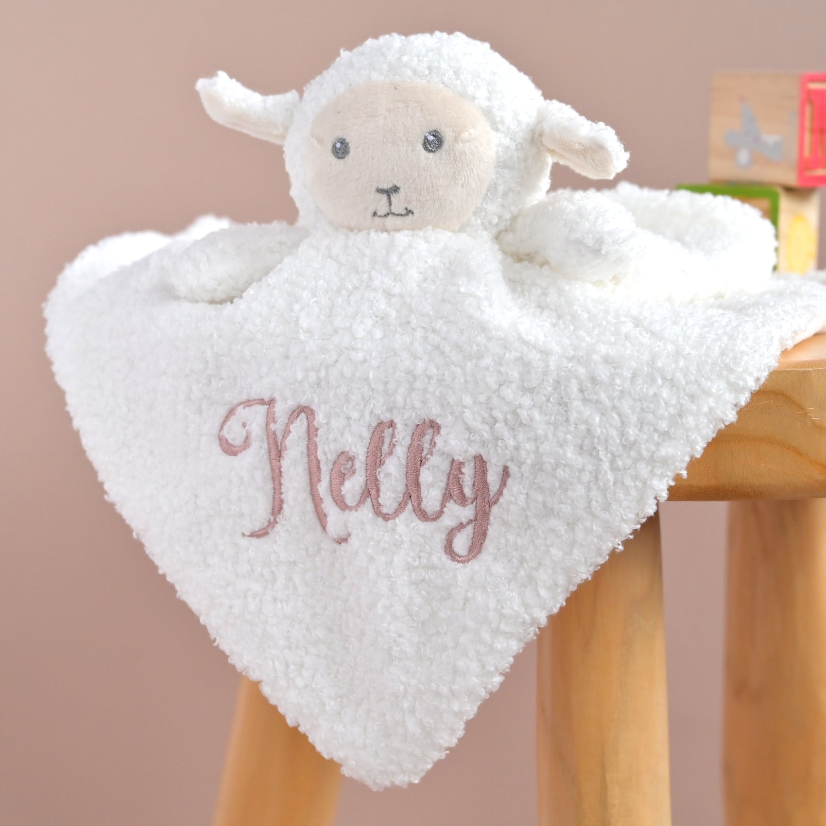 A plush white sheep lovey security blanket with the name 'Nelly' embroidered in dusty pink script, draped over a wooden stool against a soft beige background.