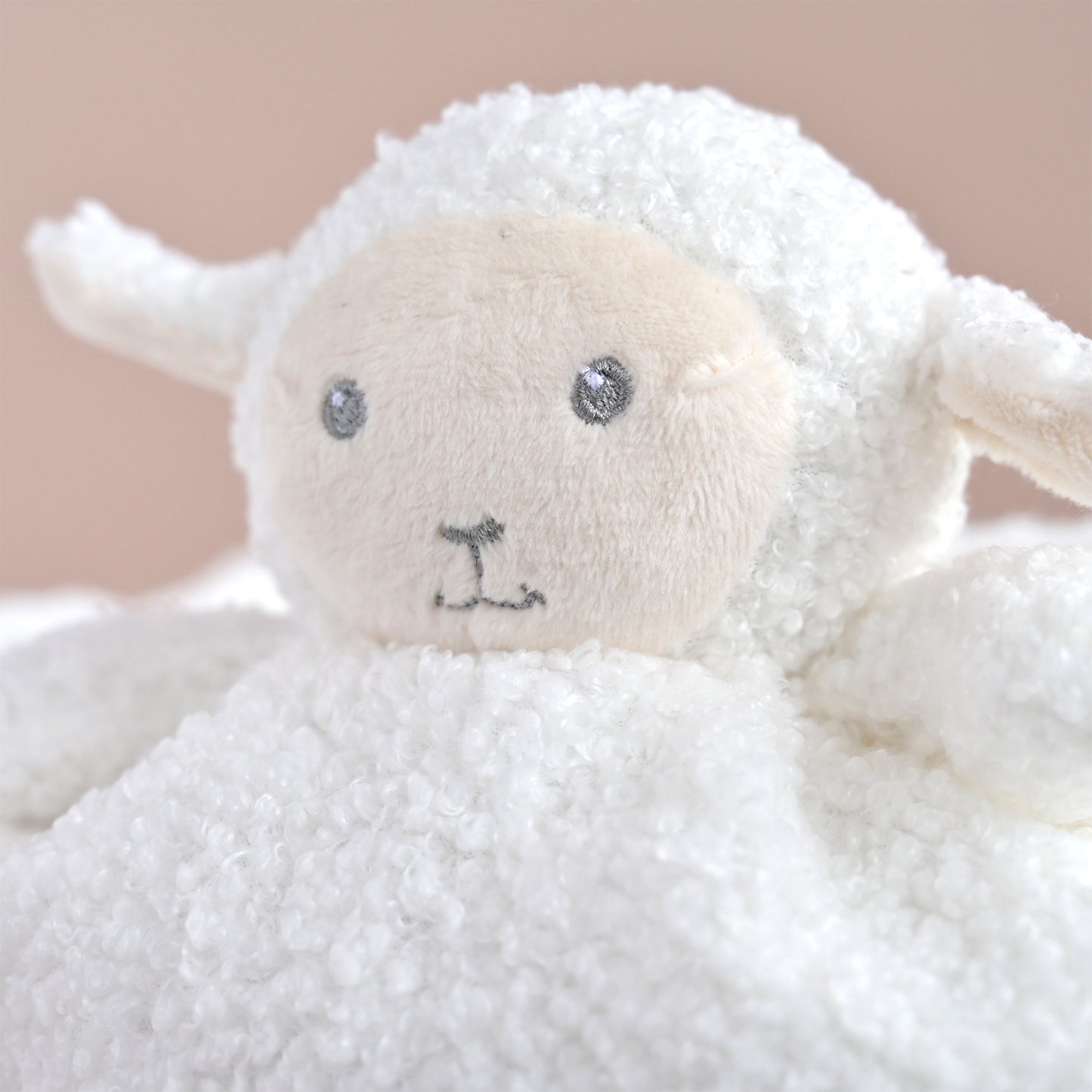 A close-up of a white plush sheep toy with a soft cream-colored face, embroidered gray eyes, and a small gray stitched nose, set against a warm beige background.