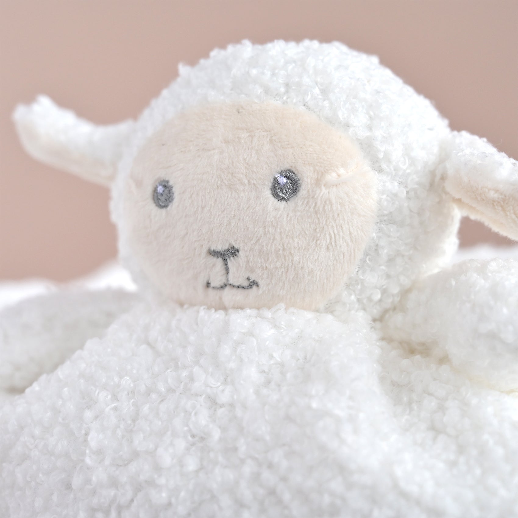 A close-up of a white plush sheep toy with a soft cream-colored face, embroidered gray eyes, and a small gray stitched nose, set against a warm beige background.