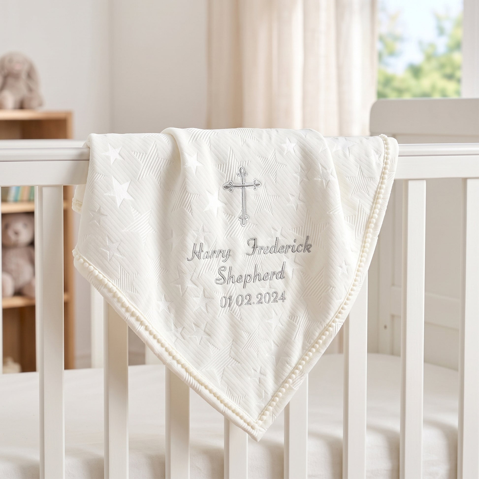 Ivory star jacquard christening blanket with pom-pom trim and scroll cross, embroidered with baby's name and date, on a cot rail