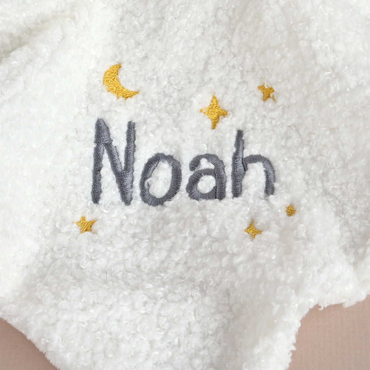 Close-up of a white sherpa fleece blanket with personalized embroidery. The name 'Noah' is stitched in gray, surrounded by small yellow stars and a yellow crescent moon.