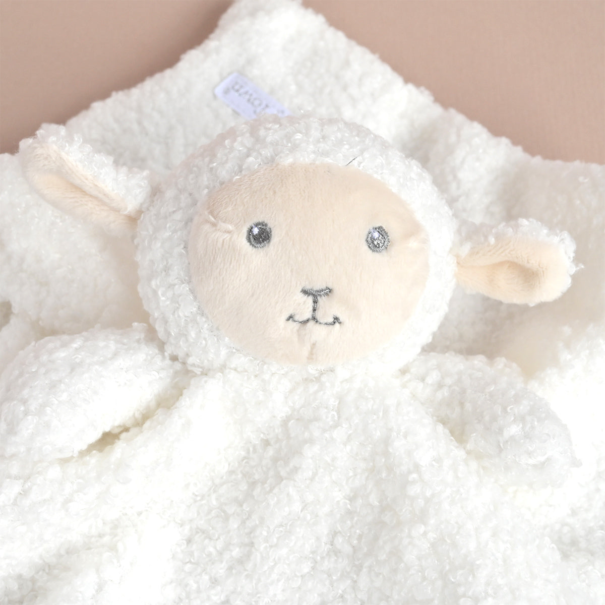 A close-up of a white fluffy sheep security blanket with a soft cream-colored face, embroidered eyes, and a small stitched nose, resting on a matching soft background.