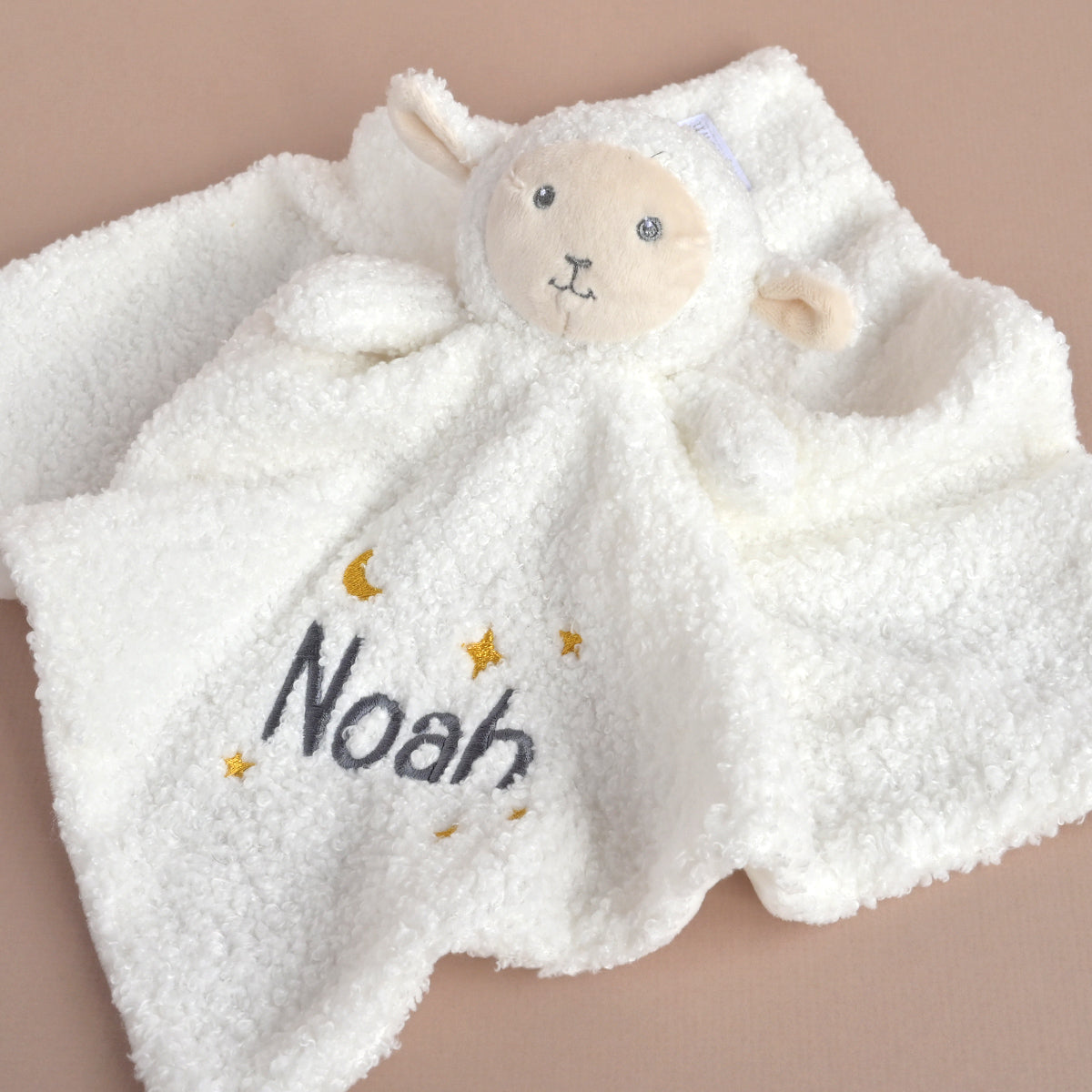 A white plush sheep lovey blanket with a soft teddy face, personalized with the name 'Noah' in dark gray embroidery and small gold stars and moon accents, laid flat on a solid beige background.