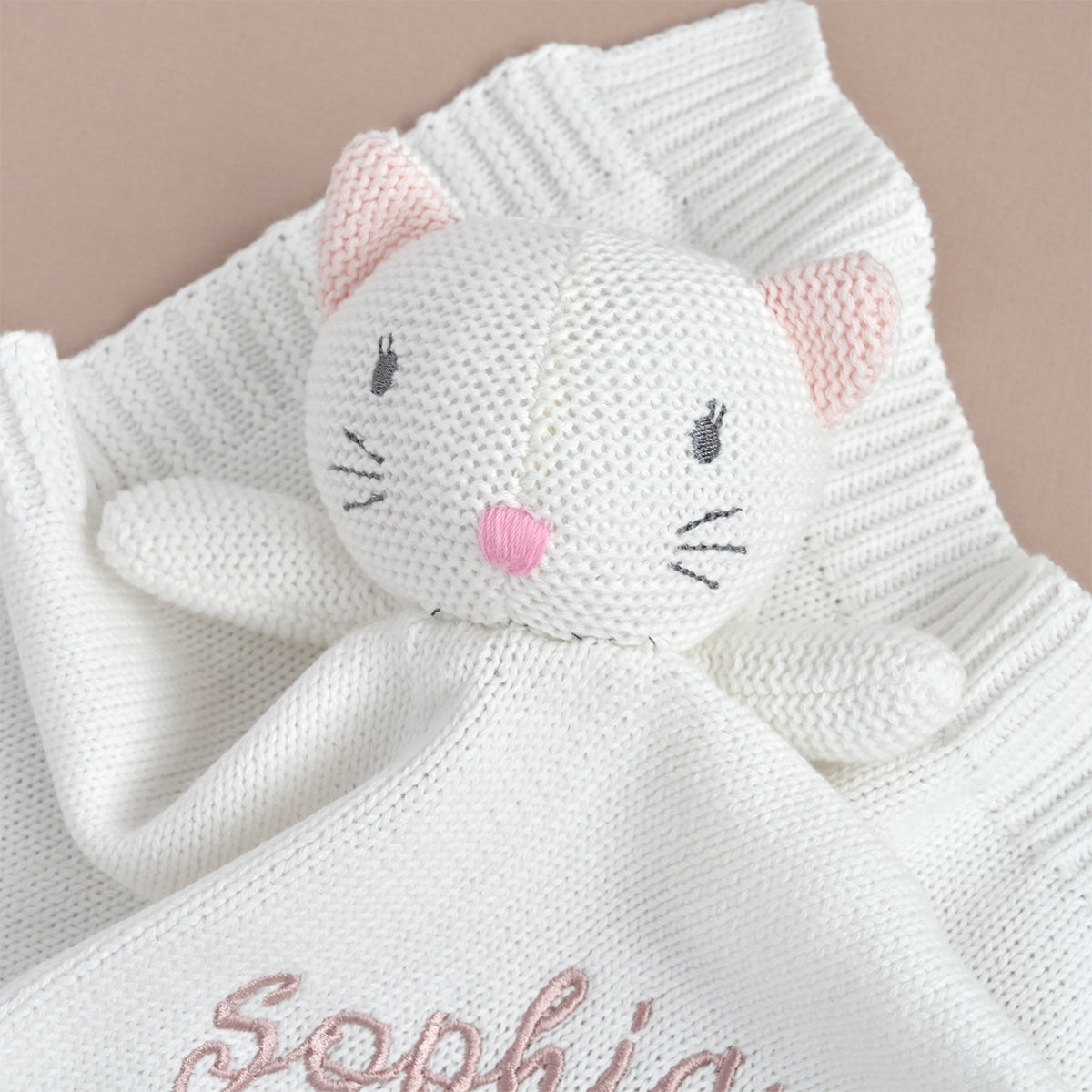 A close-up of a white knitted baby security blanket featuring a cat-shaped plush topper with pink inner ears, black embroidered eyes and whiskers, and a pink nose. The blanket is draped over a soft beige background, with the name 'Sophia' partially visible in pink script near the bottom.