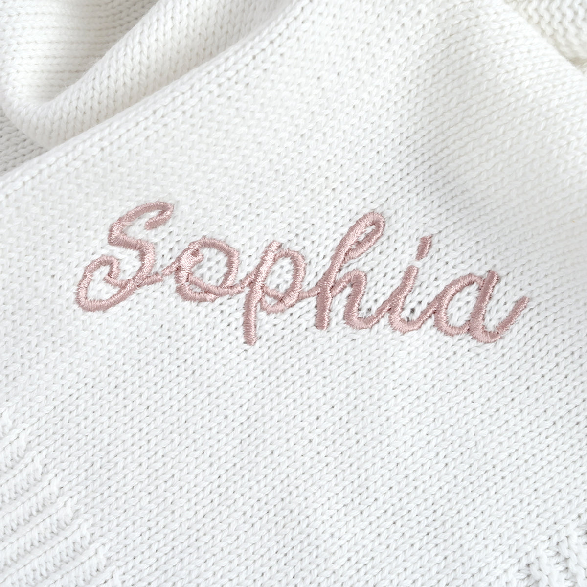 A close-up of a white knit blanket featuring the name 'Sophia' embroidered in a dusty pink script font.