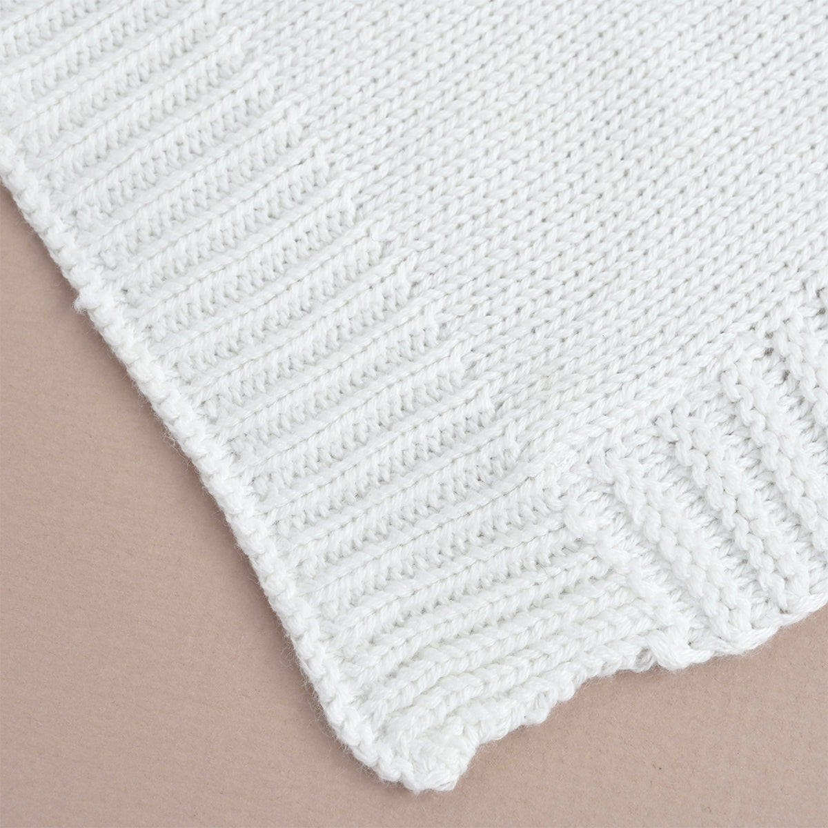 A close-up of a white knitted textile, likely a blanket or sweater corner, showing the ribbed knit texture and scalloped edge against a plain beige background.