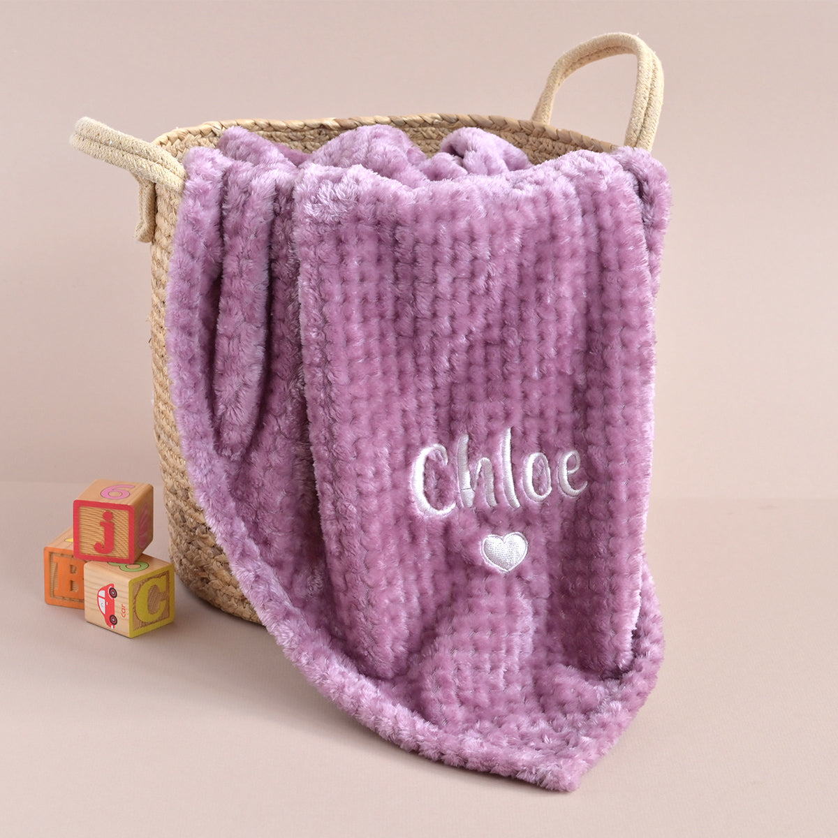 A lavender plush blanket with the name 'Chloe' and a small heart embroidered in white on the front, draped over a woven straw basket. Two small wooden blocks are placed beside the basket on a plain beige background.