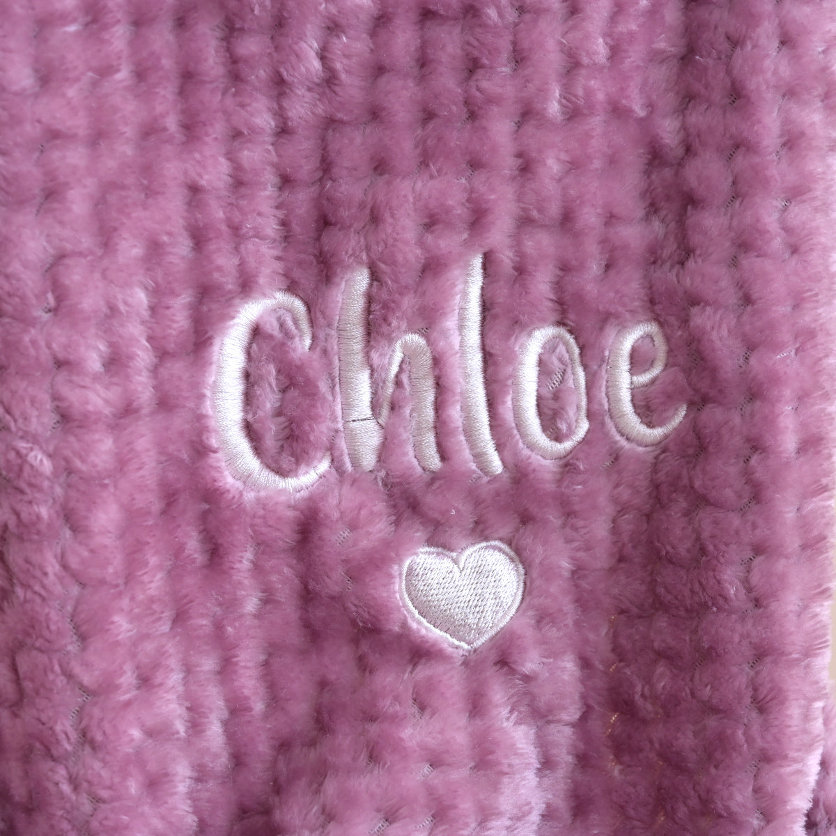 A close-up of a pink textured blanket featuring custom white embroidery with the name 'Chloe' and a small heart.