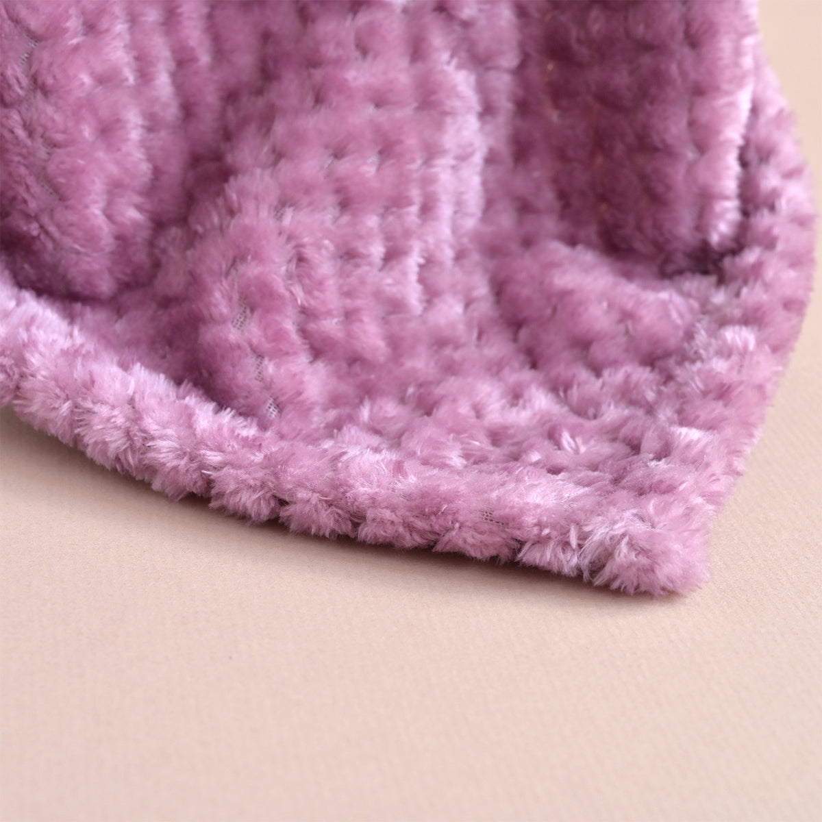 A close-up of a soft, fuzzy pink blanket or throw with a textured, plush surface, folded over to show its thick, shaggy edge against a light beige background.