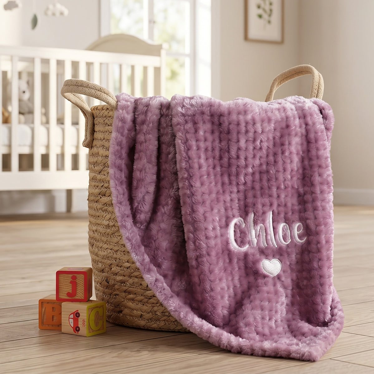 A plush lavender-purple textured blanket with the name 'Chloe' and a small white heart embroidered on it, draped over a woven straw baby basket in a softly lit nursery.