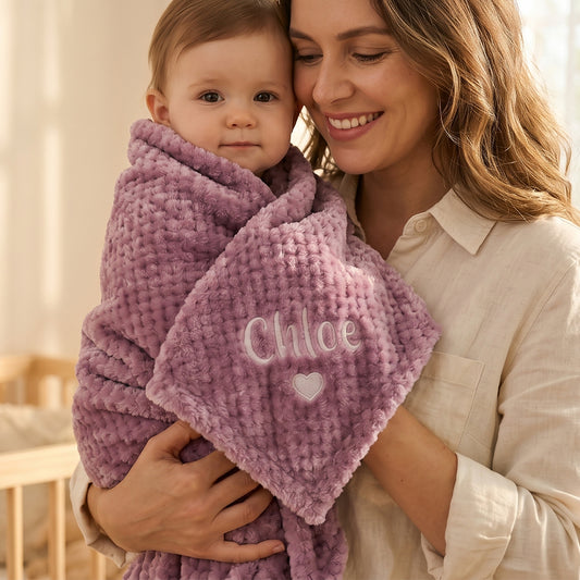 A woman in a cream shirt holds a baby wrapped in a textured lavender-purple blanket with the name 'Chloe' and a small heart embroidered on the front.