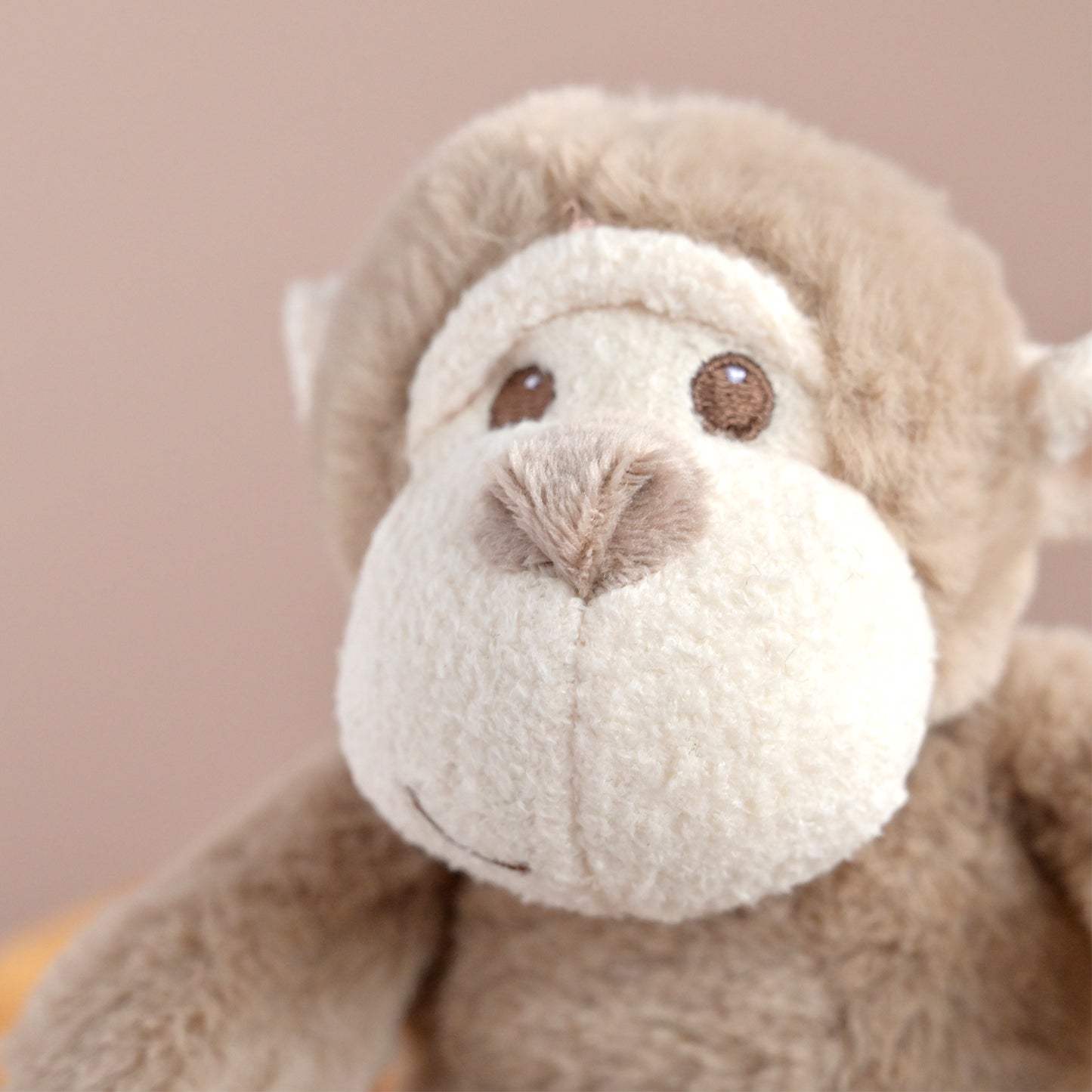 A close-up of a plush monkey toy with soft tan fur, a cream-colored face, and brown embroidered eyes and nose, set against a plain warm beige background.