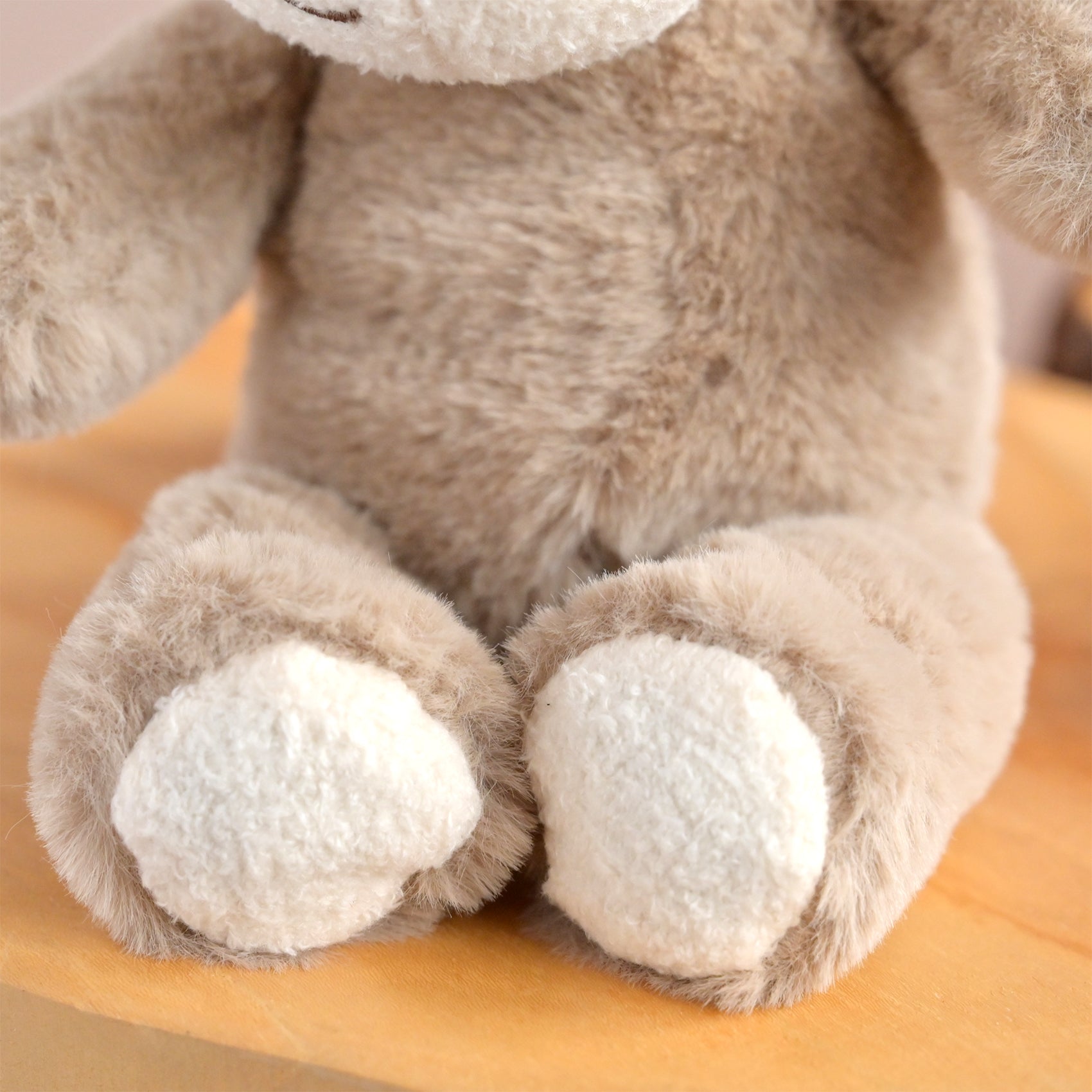 Close-up of a plush stuffed animal, showing soft beige fur, cream-colored paws, and a white muzzle on a light wooden surface.