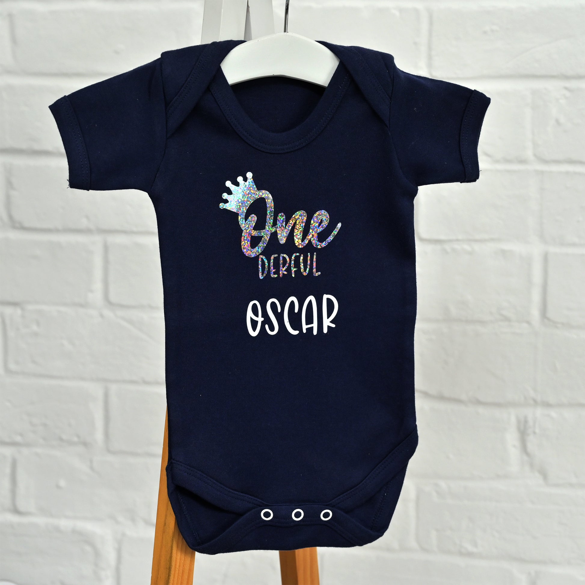 A navy blue baby bodysuit hanging on a white hanger, featuring a personalized graphic that reads 'One Derful OSCAR' with a glittery crown and lettering, set against a white brick wall background.