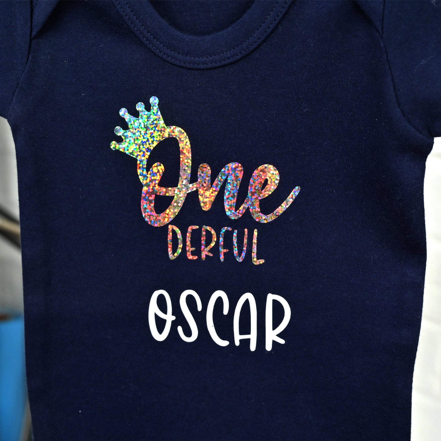 Close-up of a navy blue baby bodysuit with a colorful glittery 'One Derful' text design featuring a small crown and the name 'Oscar' in white.