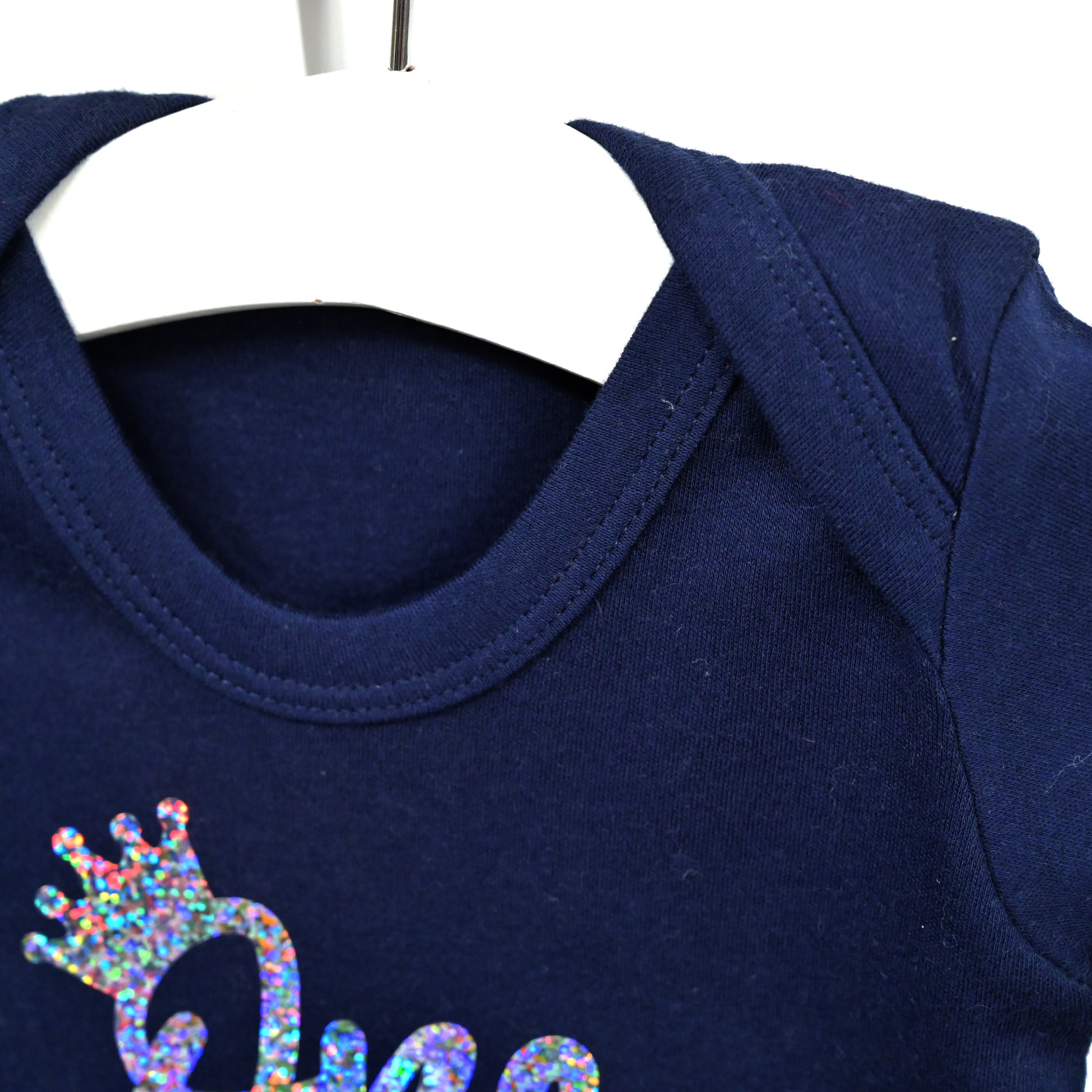 Close-up of a navy blue baby bodysuit on a white hanger, featuring a colorful holographic glitter crown graphic and the partial text 'Princ' on the chest.
