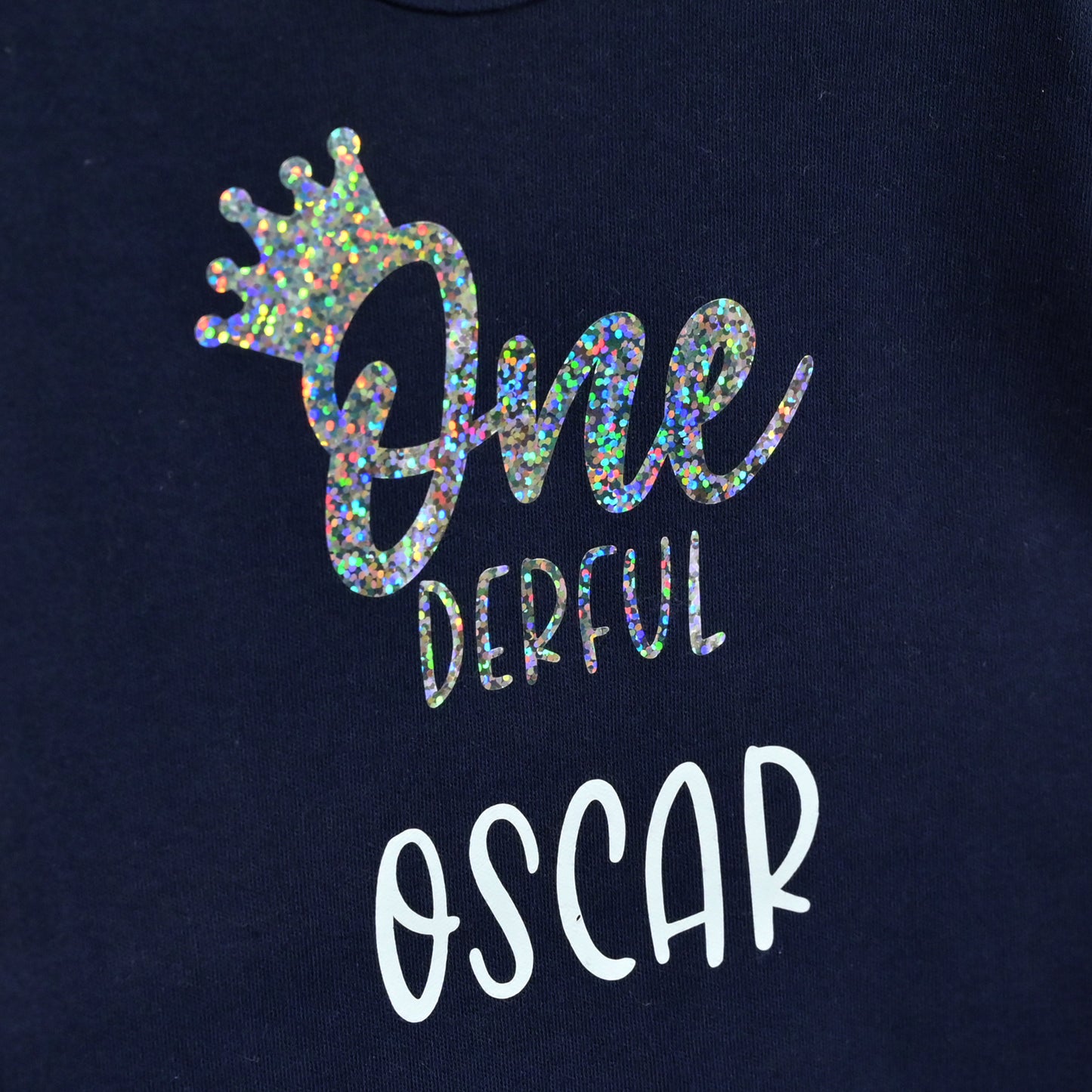 Close-up of a navy blue sweatshirt featuring a custom graphic print. The design includes a holographic glitter crown above the text 'One Derful' in a mix of holographic and white fonts, and the name 'OSCAR' printed below in white.