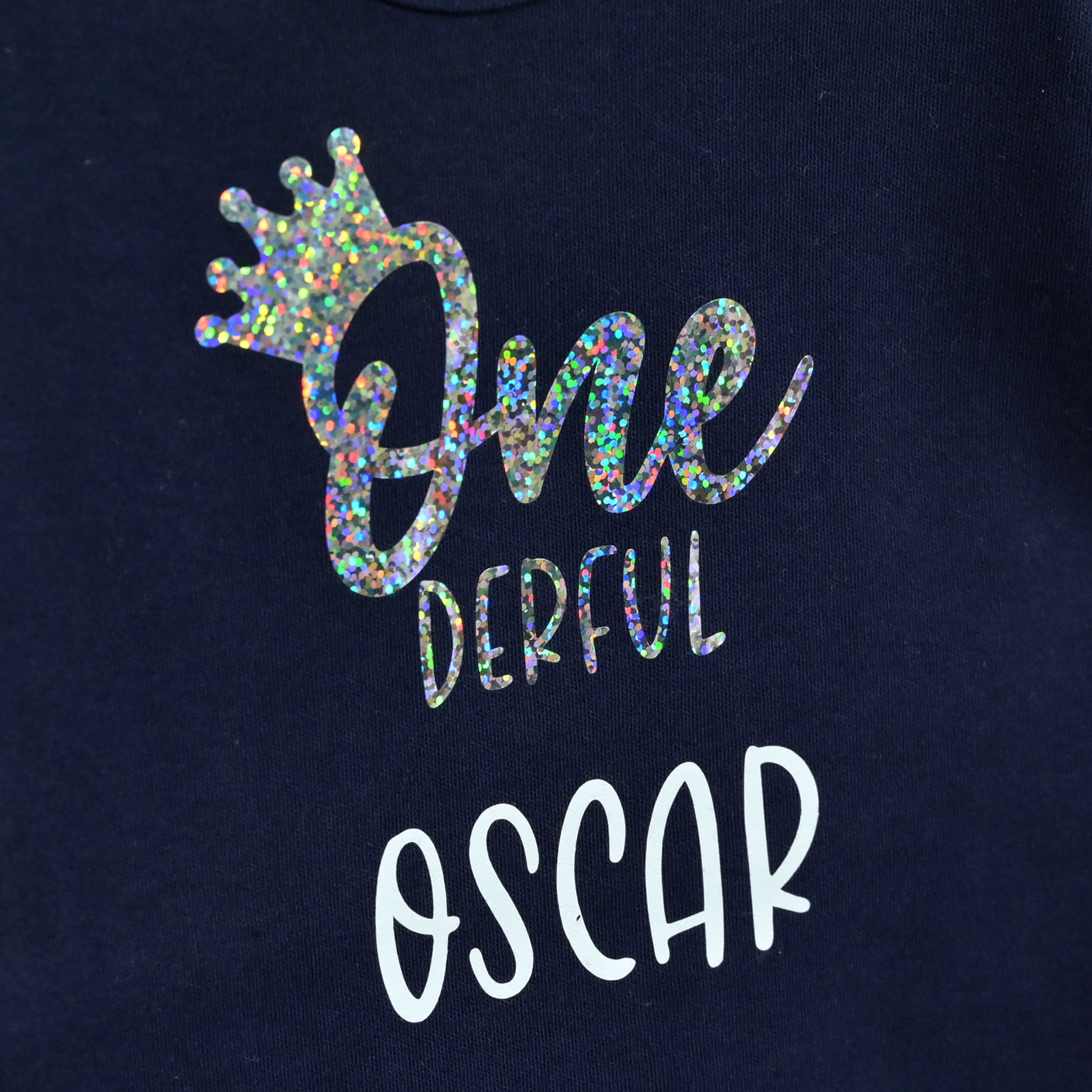 Close-up of a navy blue sweatshirt featuring a custom graphic print. The design includes a holographic glitter crown above the text 'One Derful' in a mix of holographic and white fonts, and the name 'OSCAR' printed below in white.