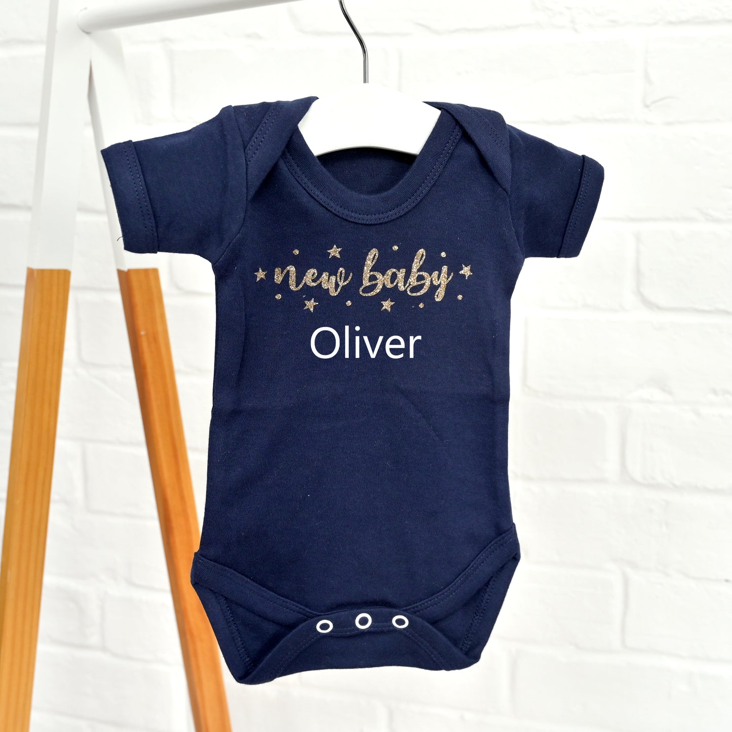 A navy blue baby bodysuit hanging on a white hanger against a white brick wall. The bodysuit features 'new baby' in gold glittery script with small gold stars, and the name 'Oliver' printed below in white.