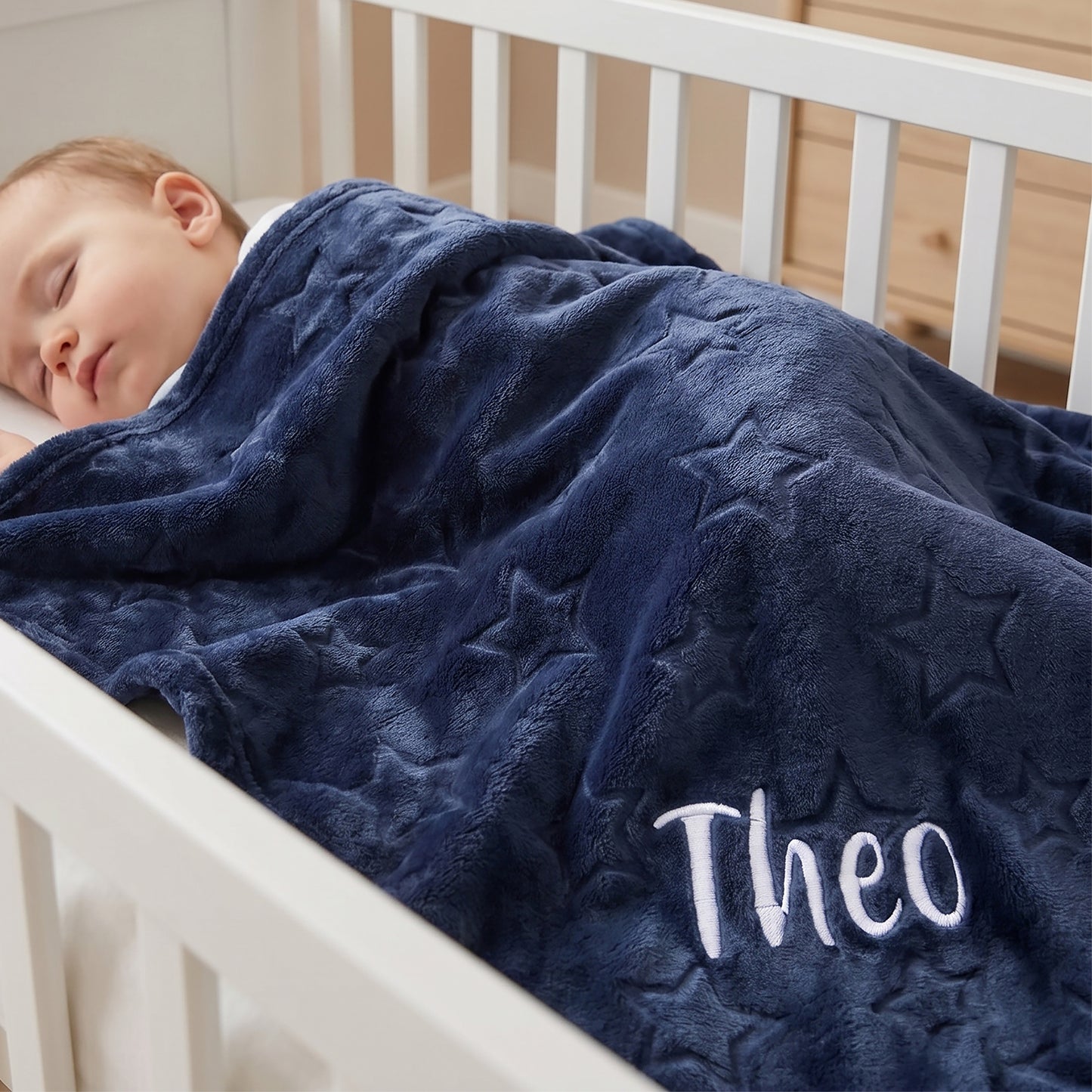 Child sleeping in a crib with a blue blanket that has 'Theo' embroidered on it.