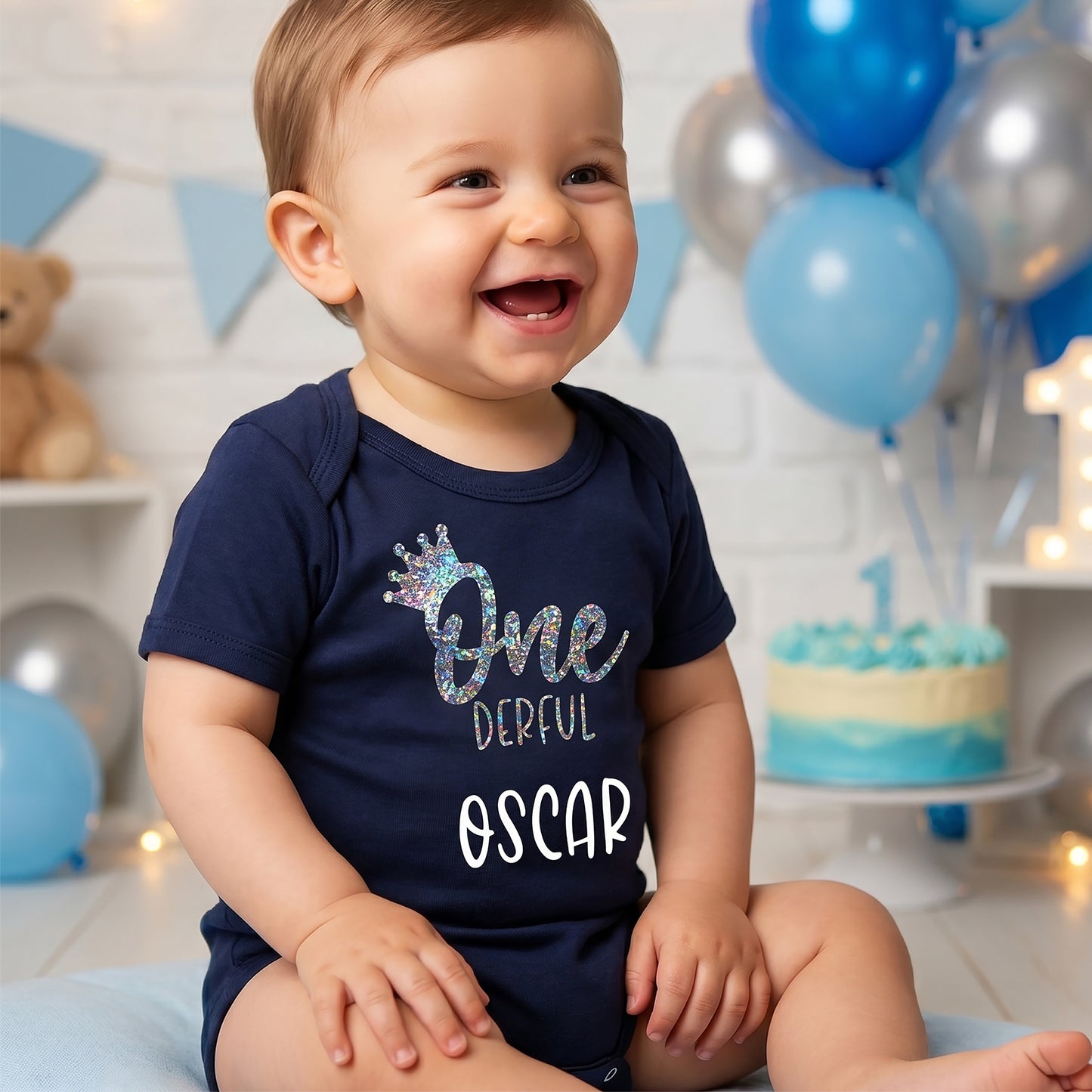 A baby wearing a navy blue short-sleeve bodysuit with a glittery 'One Dereful' crown graphic and the name 'Oscar' in white, sitting in a decorated first birthday room with blue and silver balloons and a cake.