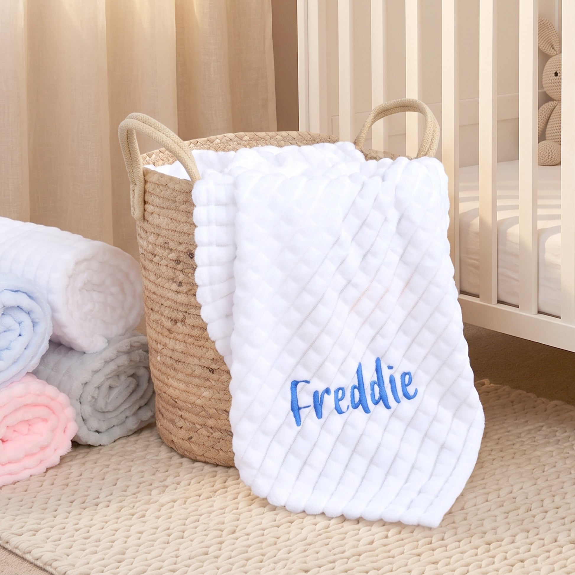White ribbed fleece baby blanket personalised with the name Freddie, folded in a wicker basket in a nursery.