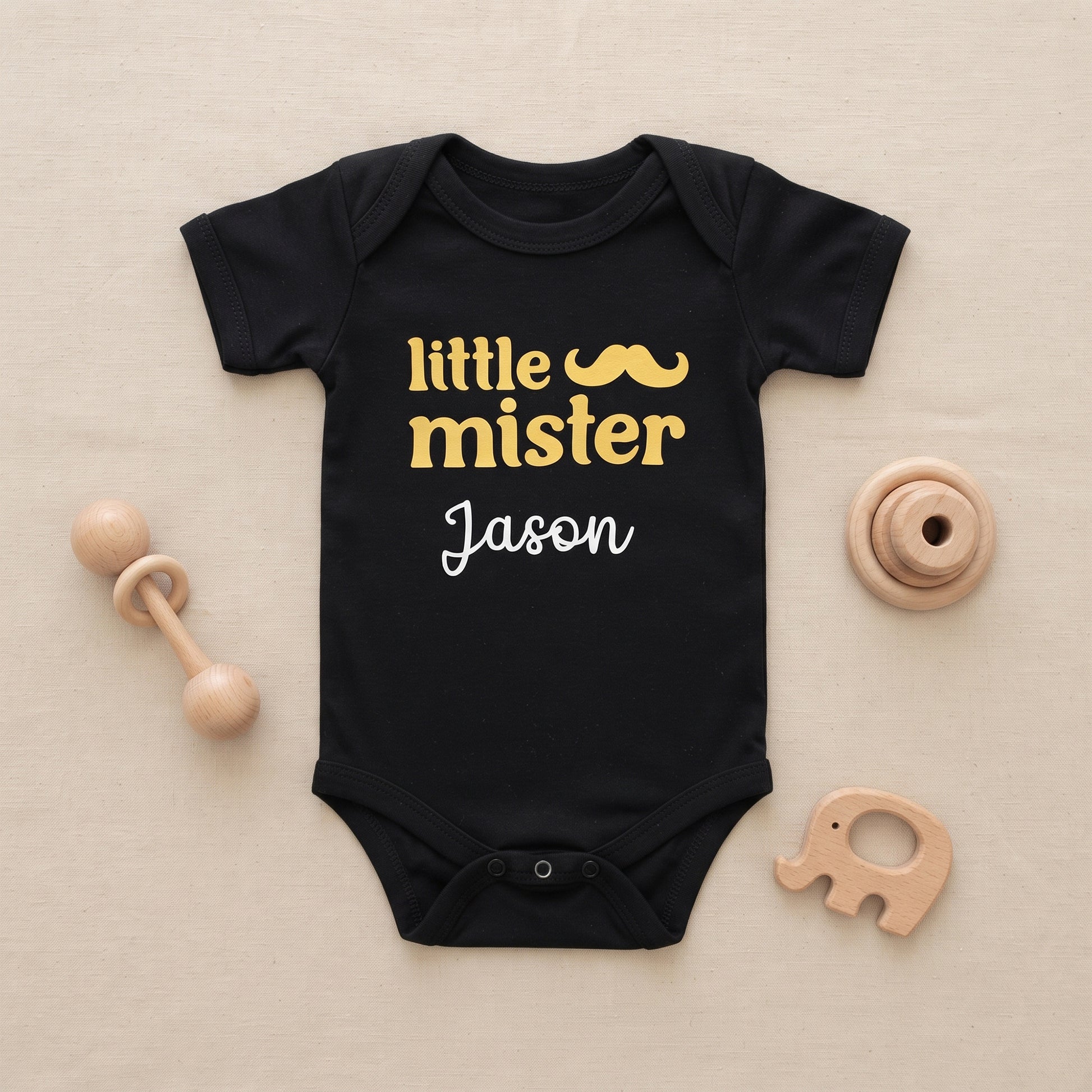 A black short-sleeve baby bodysuit laid flat on a light beige surface, printed with the words 'little mister' in yellow with a mustache graphic and 'Jason' in white script. Wooden baby toys are arranged around it.